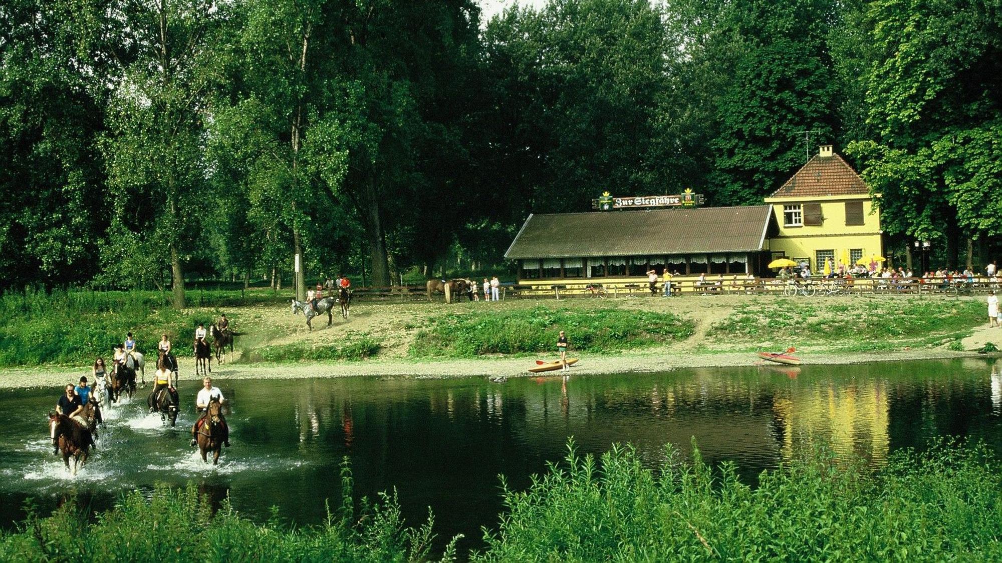 Die Aufnahme zeigt eine Gruppe, die mit Pferden die Sieg überquert. Im Hintergrund ist das Restaurant „Zur Siegfähre“ zu sehen.