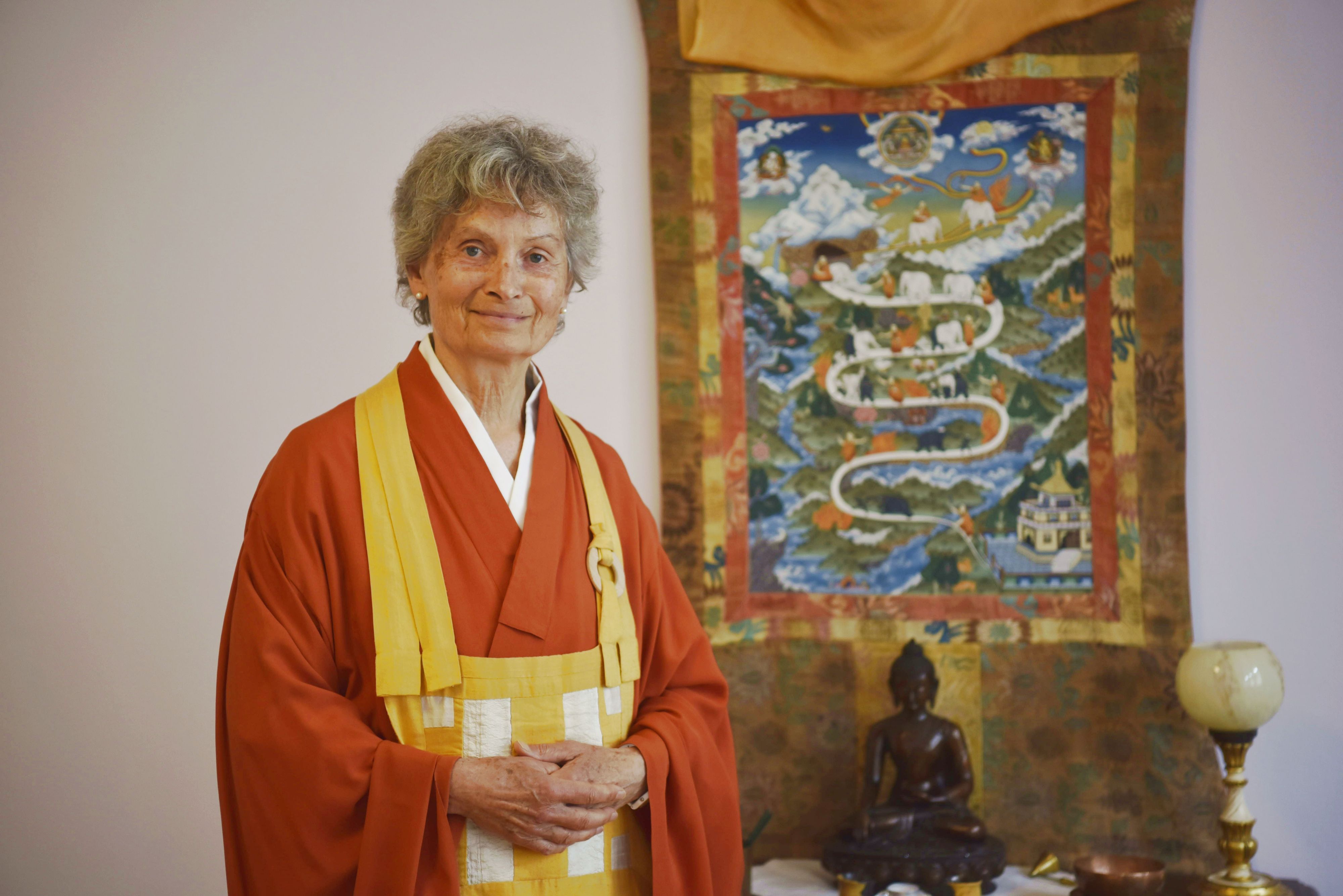 Zen-Meisterin Jigen Roshi Zeitler steht in orangenem Gewand vor einem Wandbehang und einer Buddha-Statue.