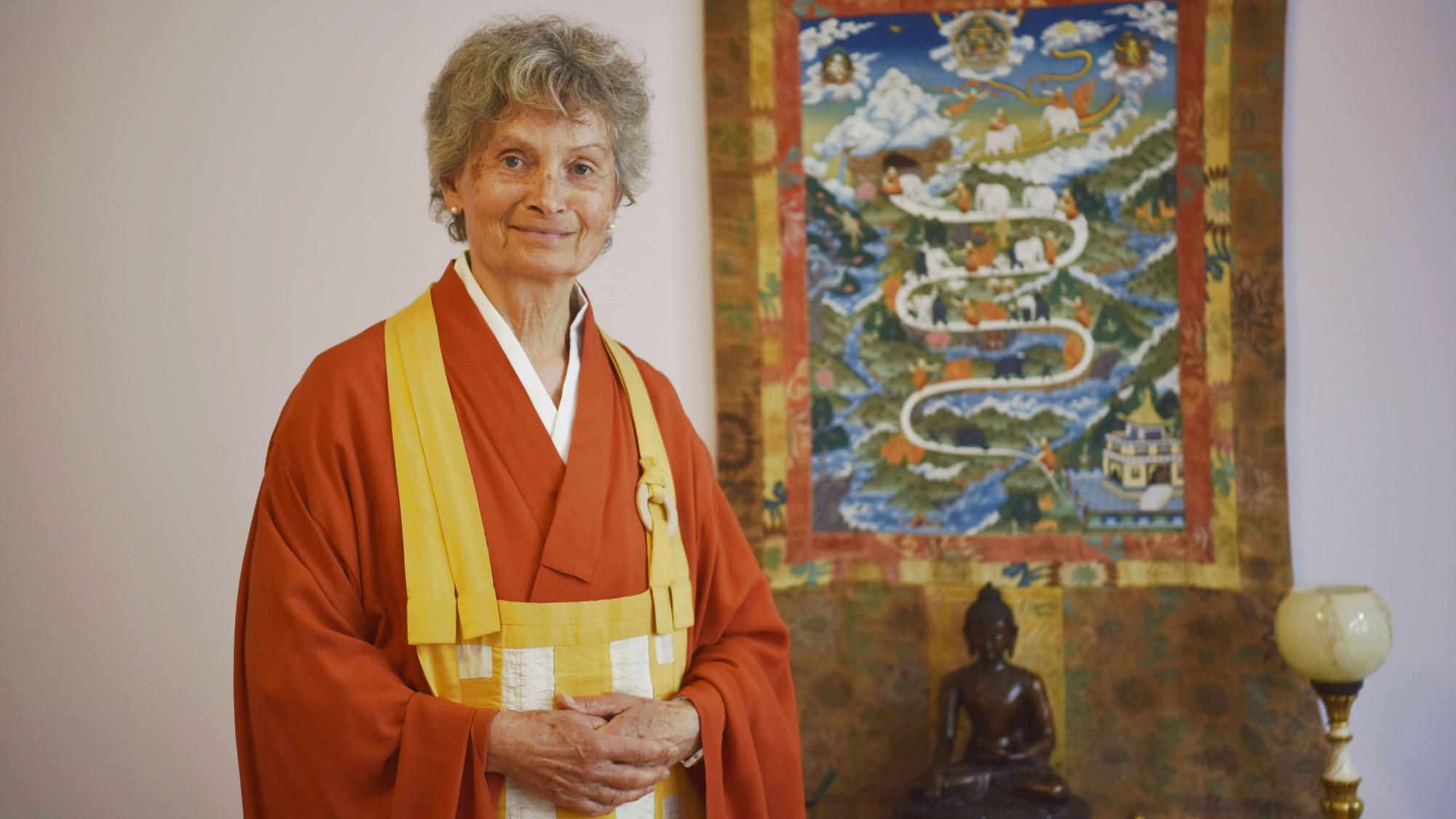 Zen-Meisterin Jigen Roshi Zeitler steht in orangenem Gewand vor einem Wandbehang und einer Buddha-Statue.