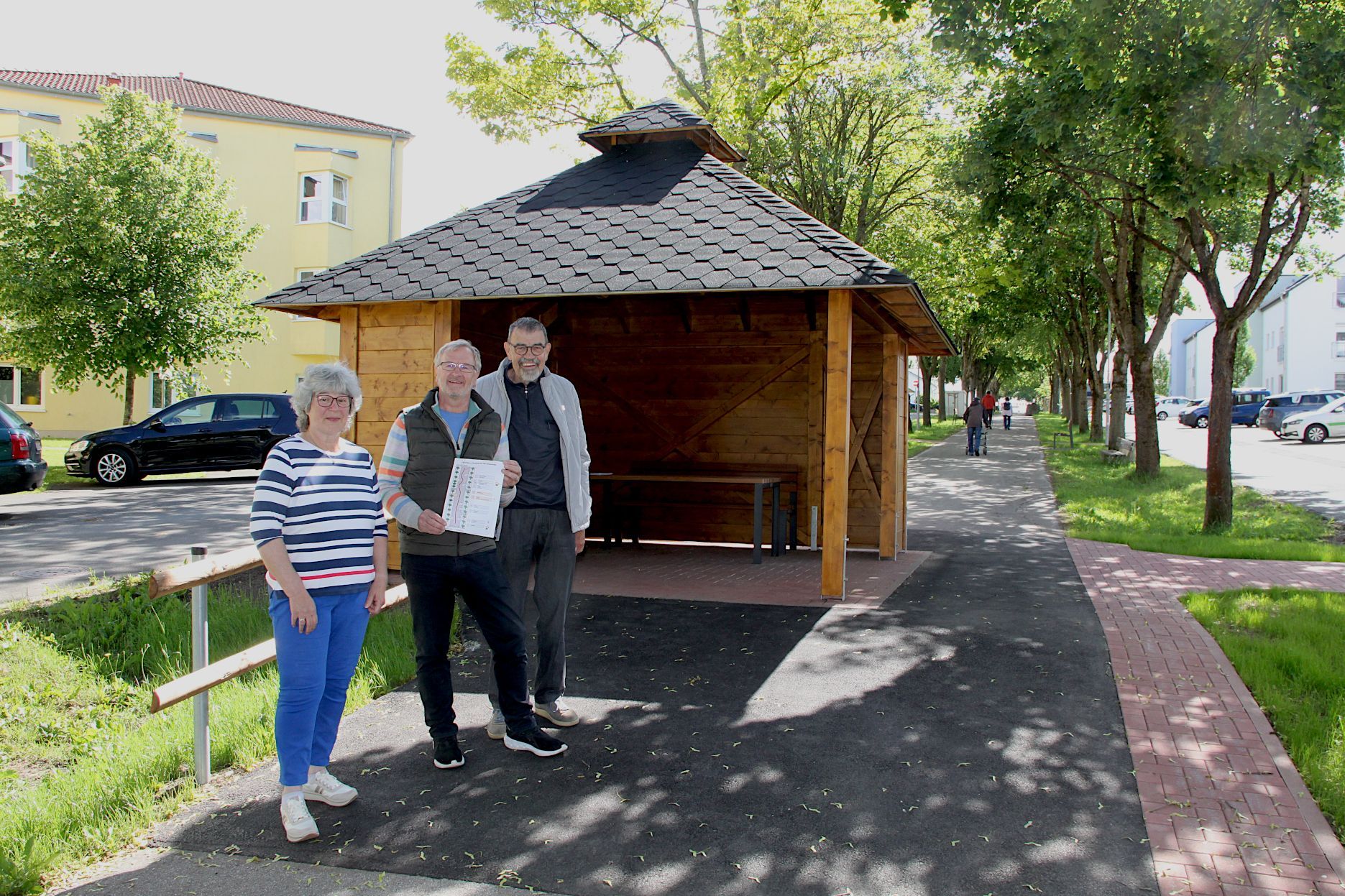 Eine Frau und zwei Männer stehen vor einem Pavillion, rundherum sind Bäume und Häuser zu erkennen.