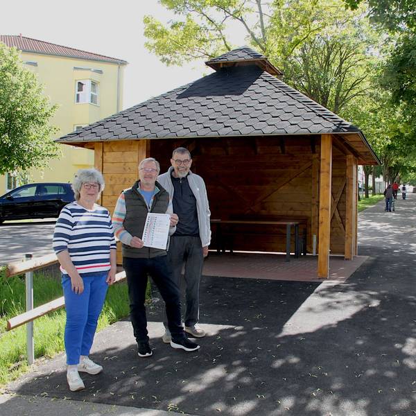 Eine Frau und zwei Männer stehen vor einem Pavillion, rundherum sind Bäume und Häuser zu erkennen.