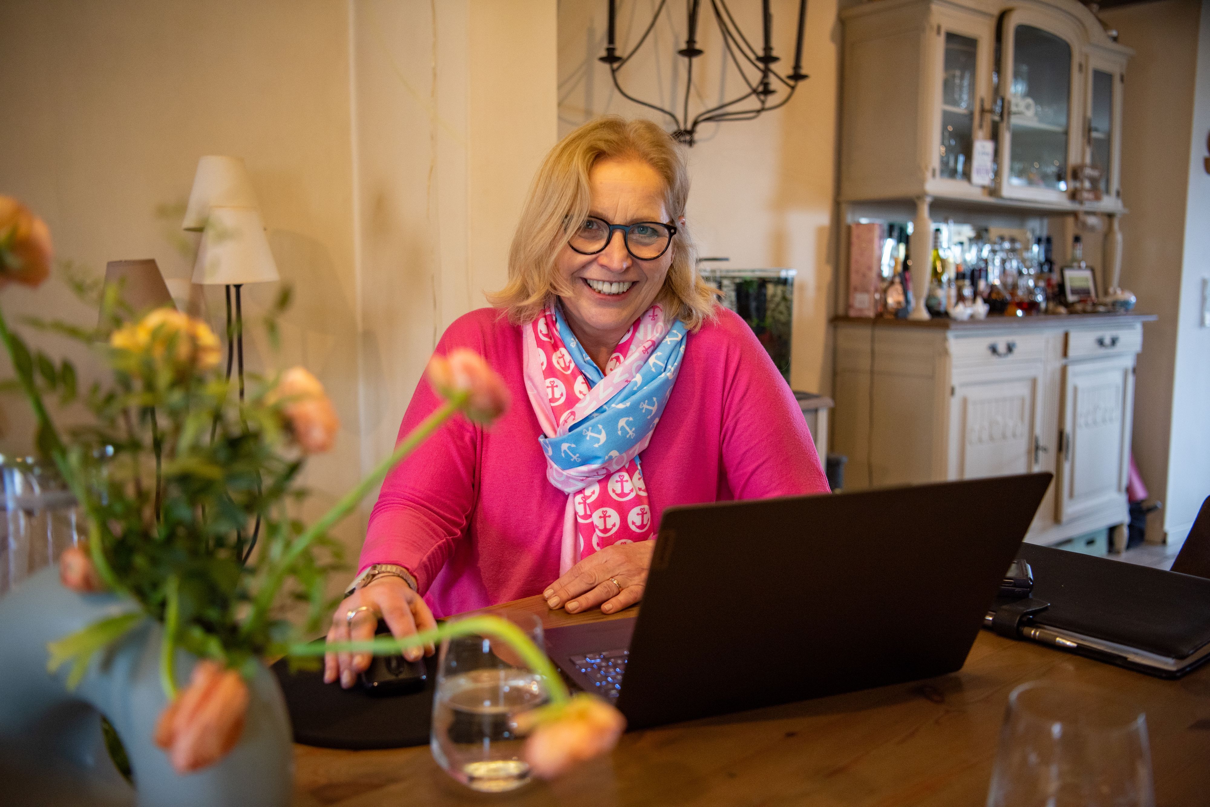 Die freie Rednerin Isa Oestmann sitzt an ihrem  Esstisch vor einem Laptop.  Vor ihr auf dem Tisch steht ein Strauß Blumen. Sie trägt ein pinkes Sweatshirt und einen Schal.