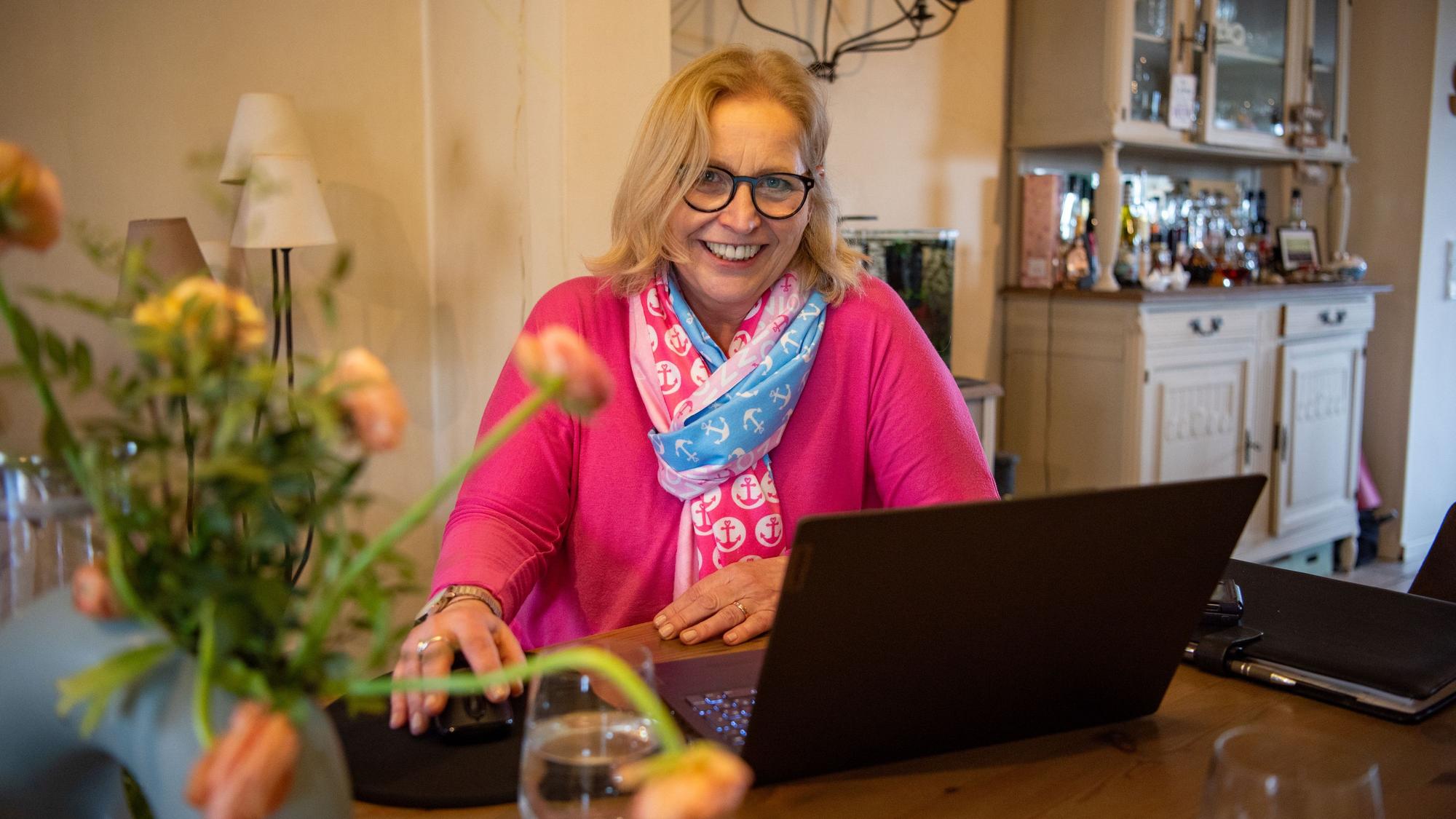 Die freie Rednerin Isa Oestmann sitzt an ihrem Esstisch vor einem Laptop. Vor ihr auf dem Tisch steht ein Strauß Blumen. Sie trägt ein pinkes Sweatshirt und einen Schal.