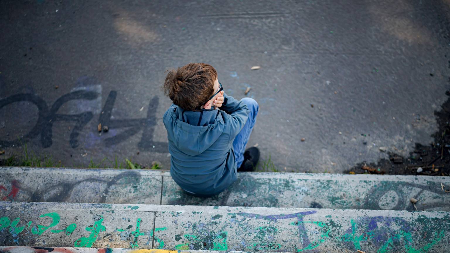 Ein Junge sitzt alleine auf einem mit Graffiti besprühten, heruntergekommenen Spielplatz.