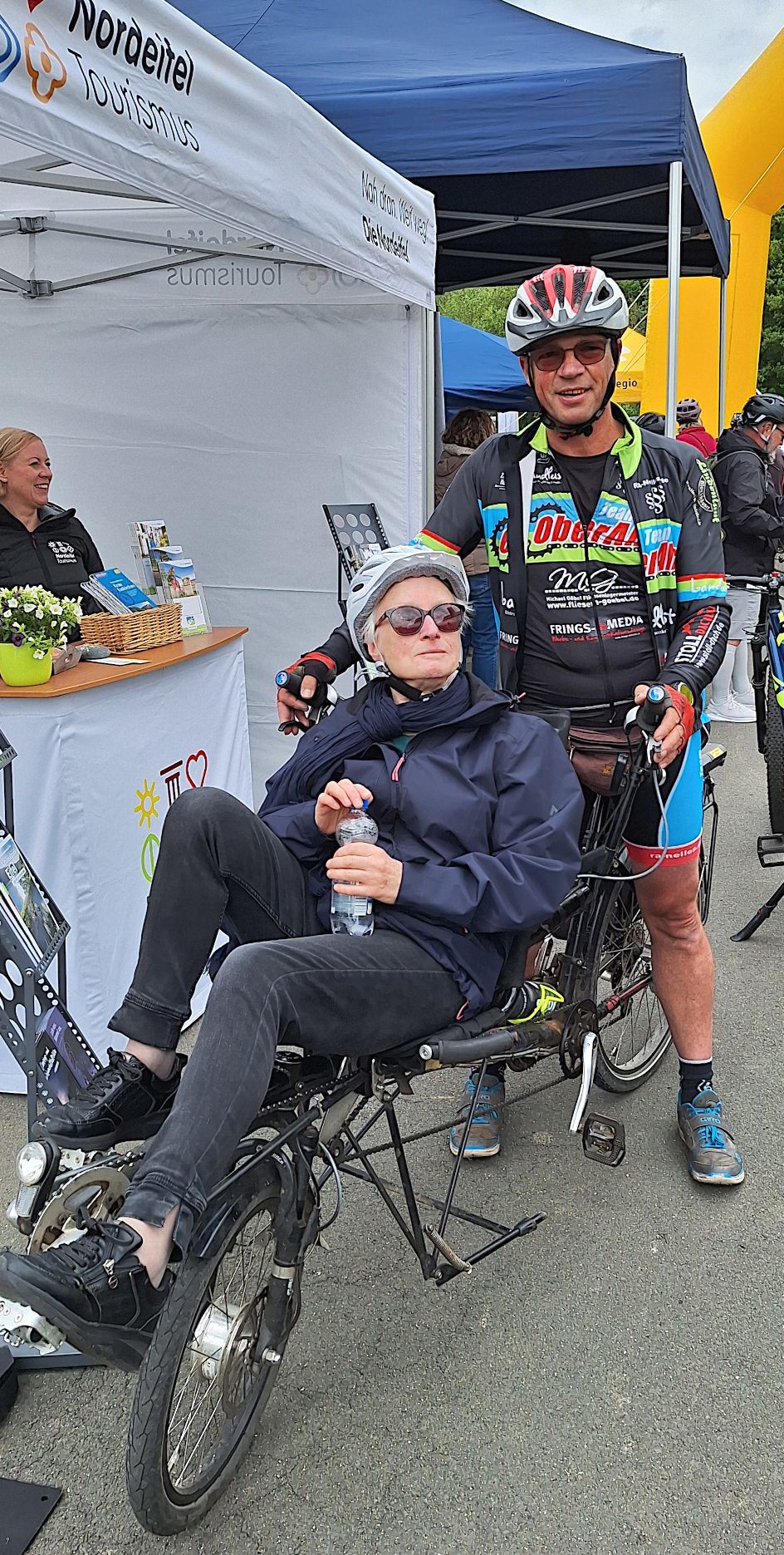 Eine Frau sitzt auf einem Sitz-Fahrrad, dahinter steht ein Mann in Radkleidung.