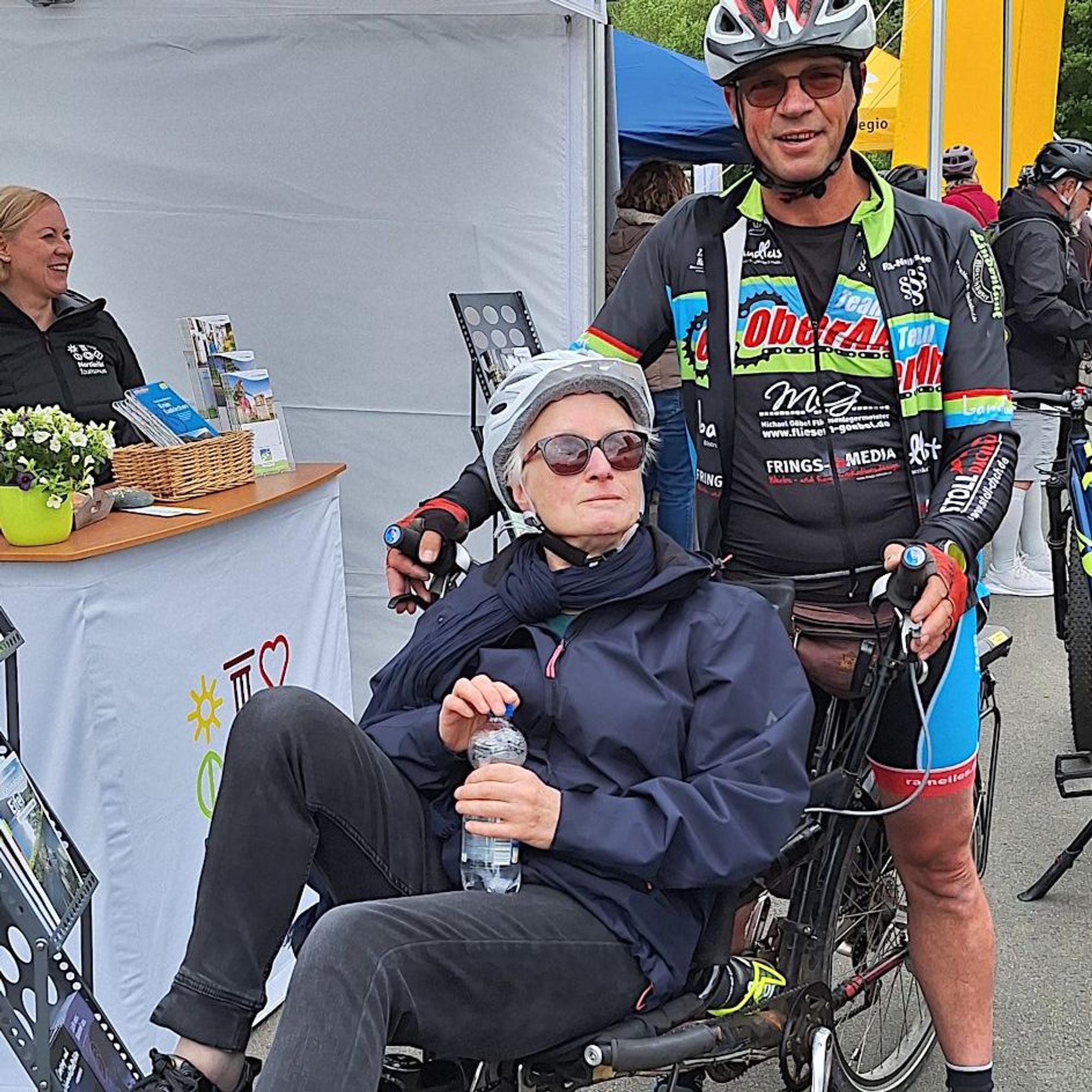 Eine Frau sitzt auf einem Sitz-Fahrrad, dahinter steht ein Mann in Radkleidung.