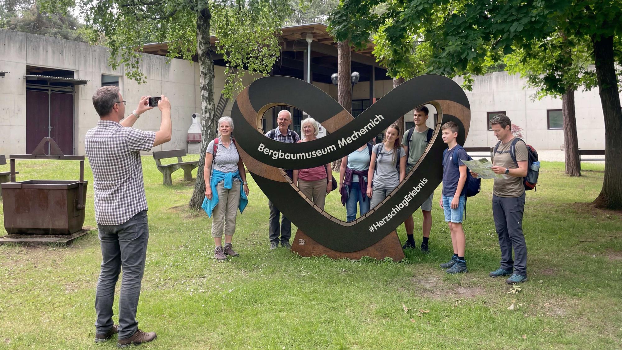 Menschen stehen um ein herförmiges Schild vor dem Bergbaumuseum in Mechernich und werden fotografiert.
