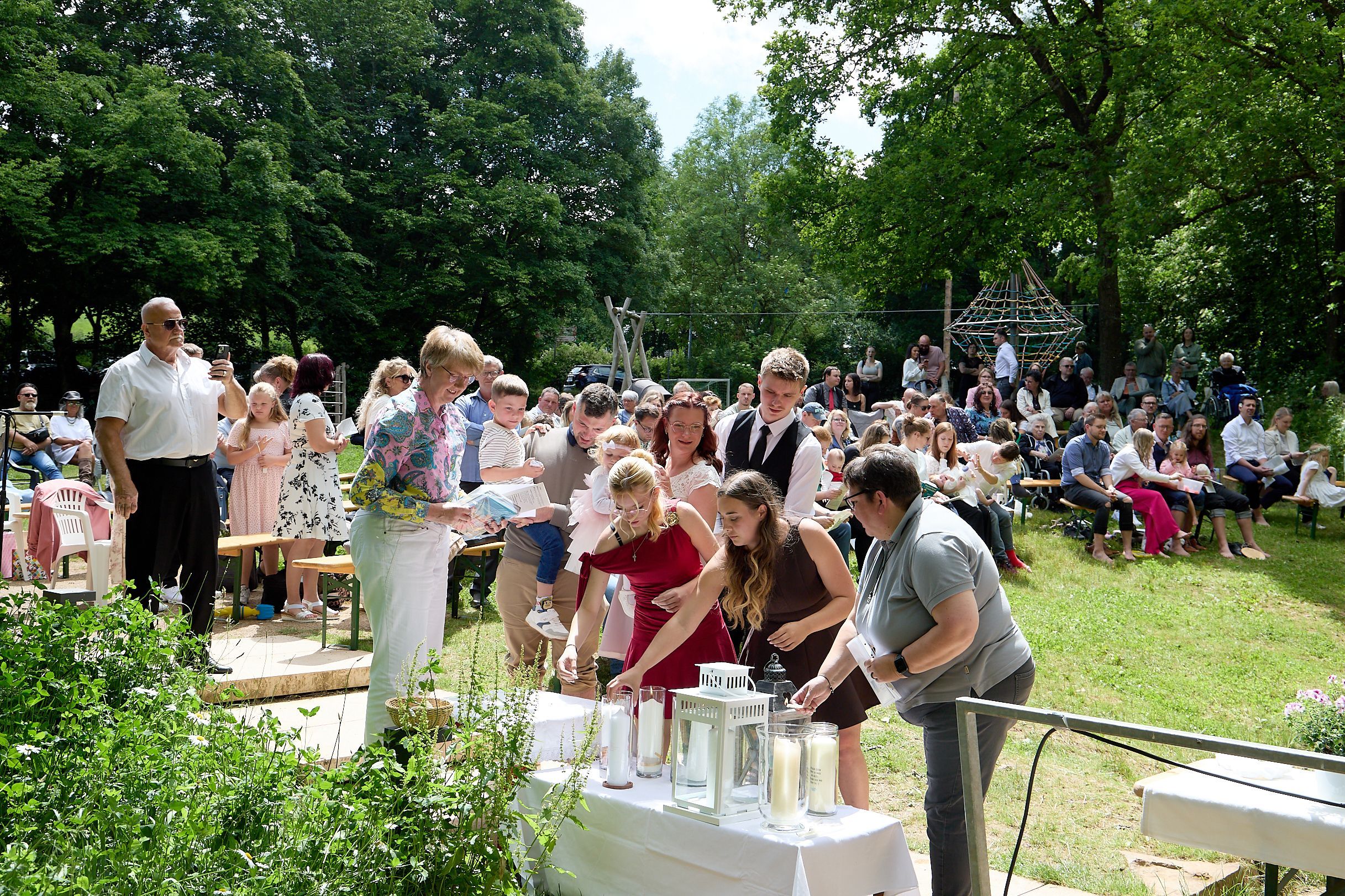 Mehrere Frauen und Männer zünden Kerzen an, die auf einem Tisch mit weißer Tischdecke stehen. Im Hintergrund sind viele weitere Menschen und Bäume zu sehen.