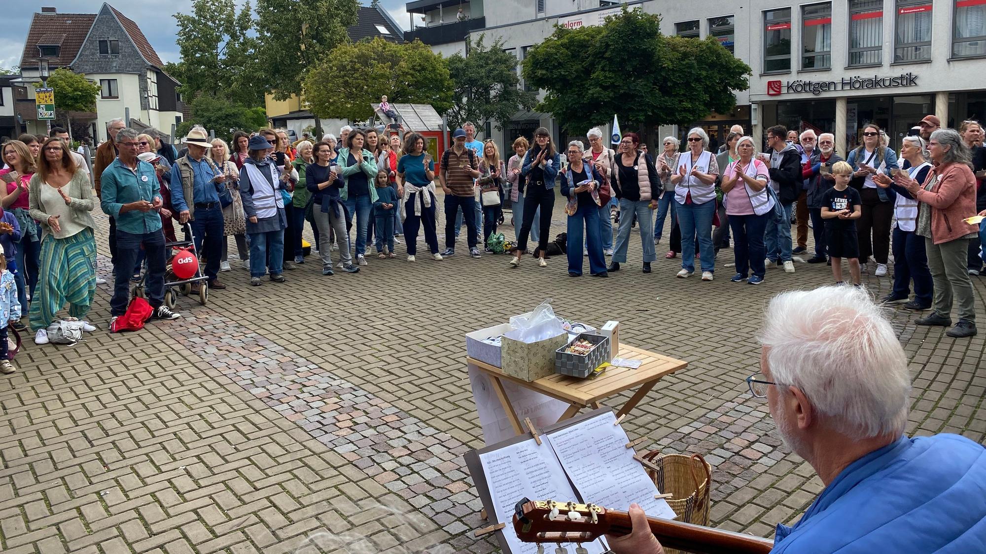 Zu sehen sind Menschen, die sich im Halbkreis aufgestellt haben. Im Vordergrund sitzt ein Mann mit einer Gitarre.