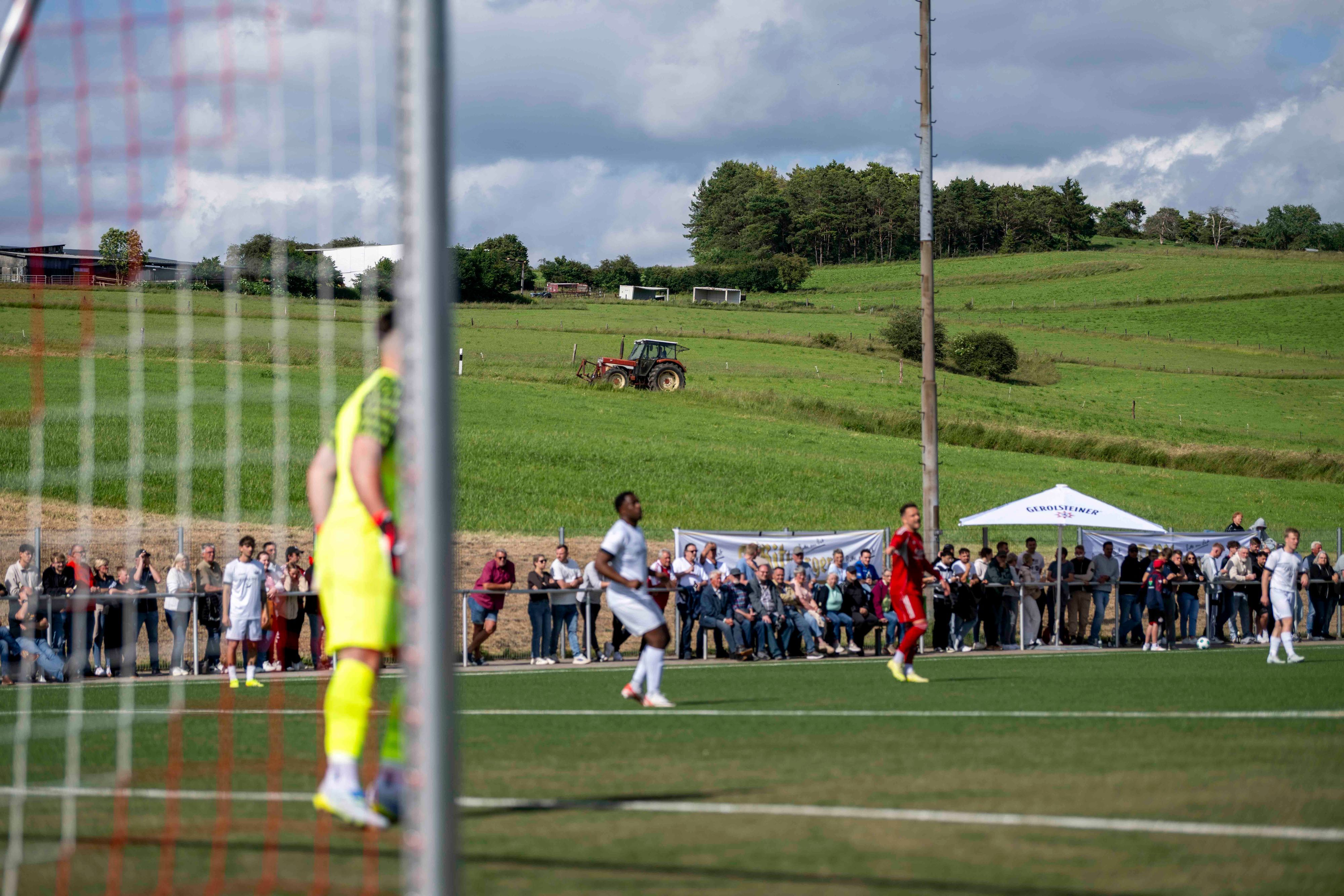 Das Bild zeigt einen Traktor, der im Hintergrund fährt. Im Vordergrund läuft das Pokalfinale zwischen Zülpich und Bessenich in Ripsdorf.