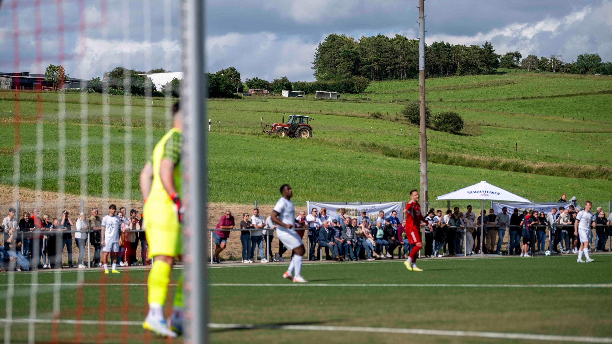 Das Bild zeigt einen Traktor, der im Hintergrund fährt. Im Vordergrund läuft das Pokalfinale zwischen Zülpich und Bessenich in Ripsdorf.