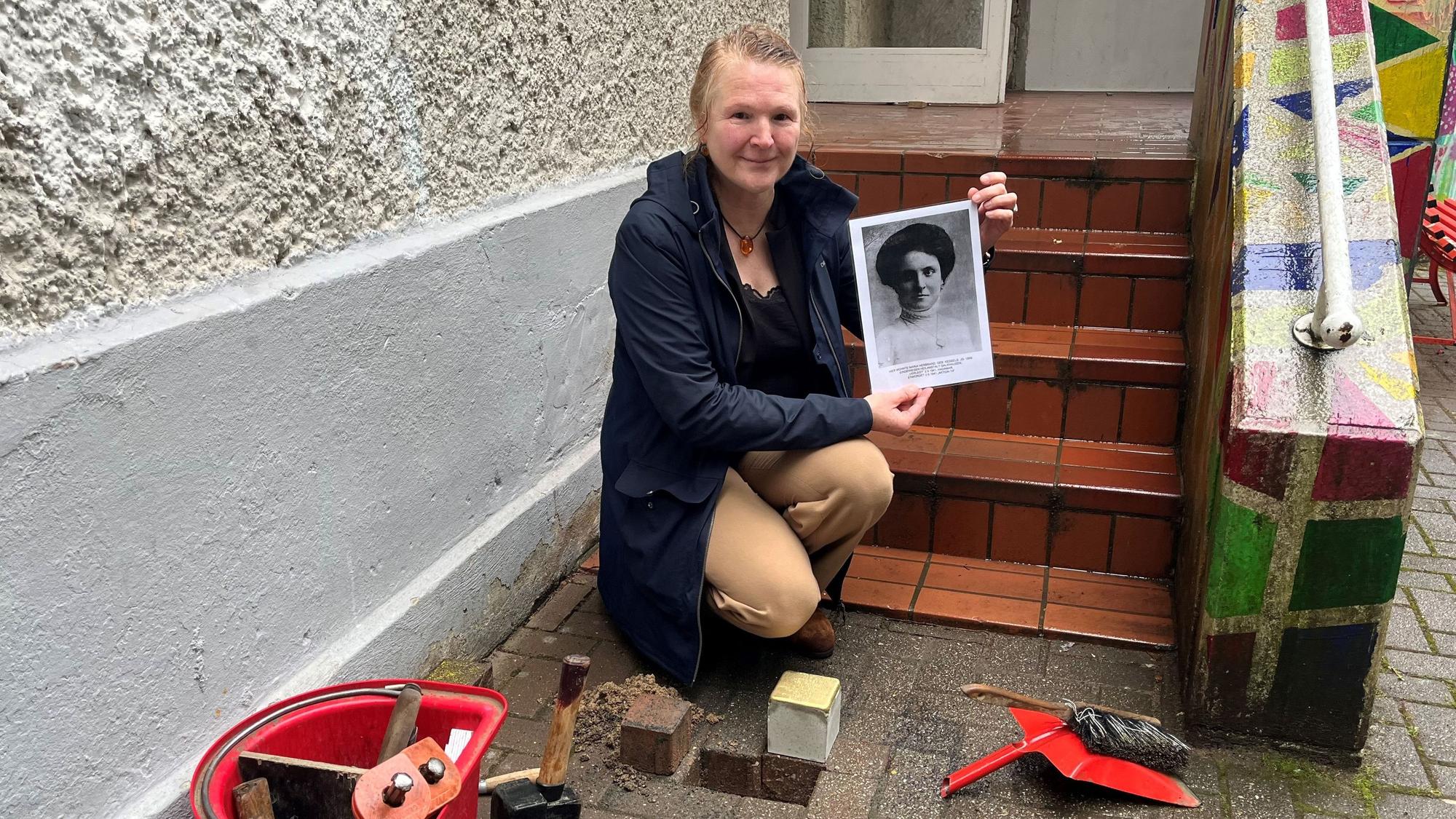 Eine Frau hockt mit einem schwarz-weißen Foto einer Frau vor einem Loch, in dem der Stolperstein verlegt werden soll.