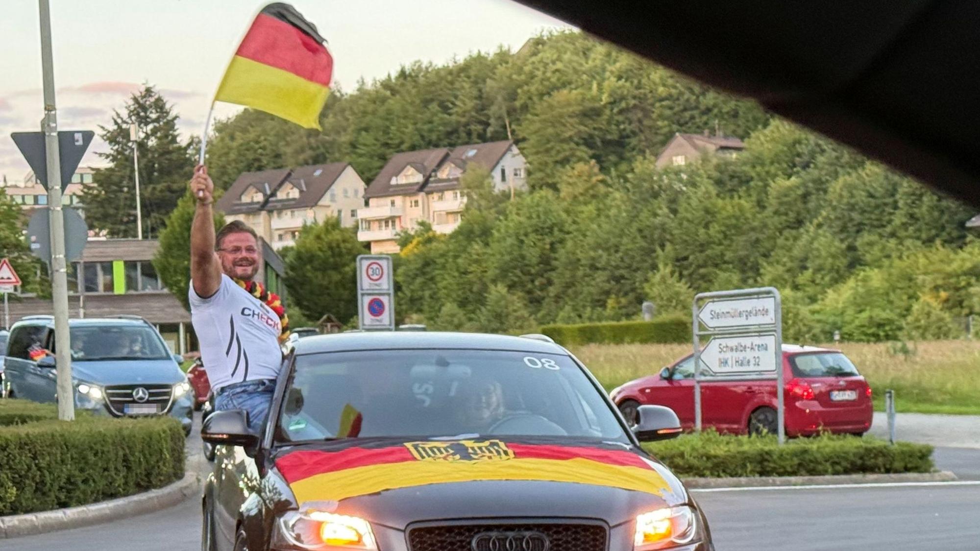 Das Foto zeigt einen Pkw. Über die Kühlerhaube ist die schwarz-rot-goldene Fahne mit Bundesadler gespannt. Der Beifahrer schwenkt die deutsche Fahne aus dem Fenster.