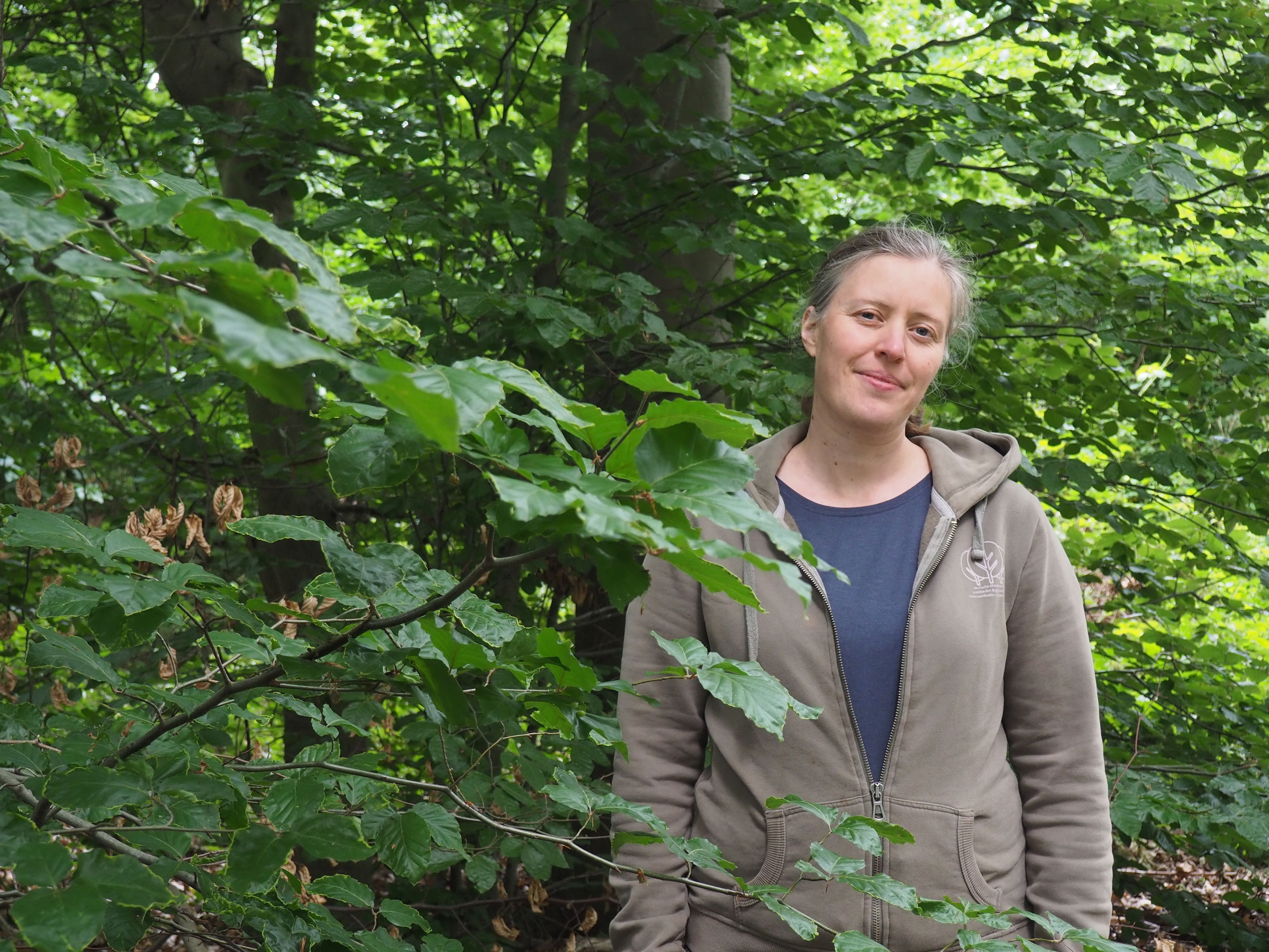 Katinka Richter trägt ein blaues Shirt und eine Kapuzenjacke und steht in einem Wald.