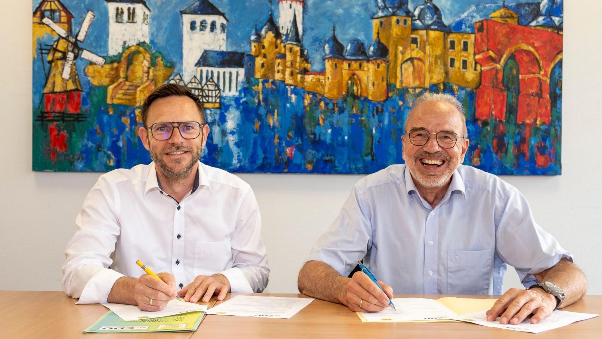 Björn Wassong und Dr. Manfred Rechs sitzen an einem Tisch und unterzeichnen eine Vereinbarung.
