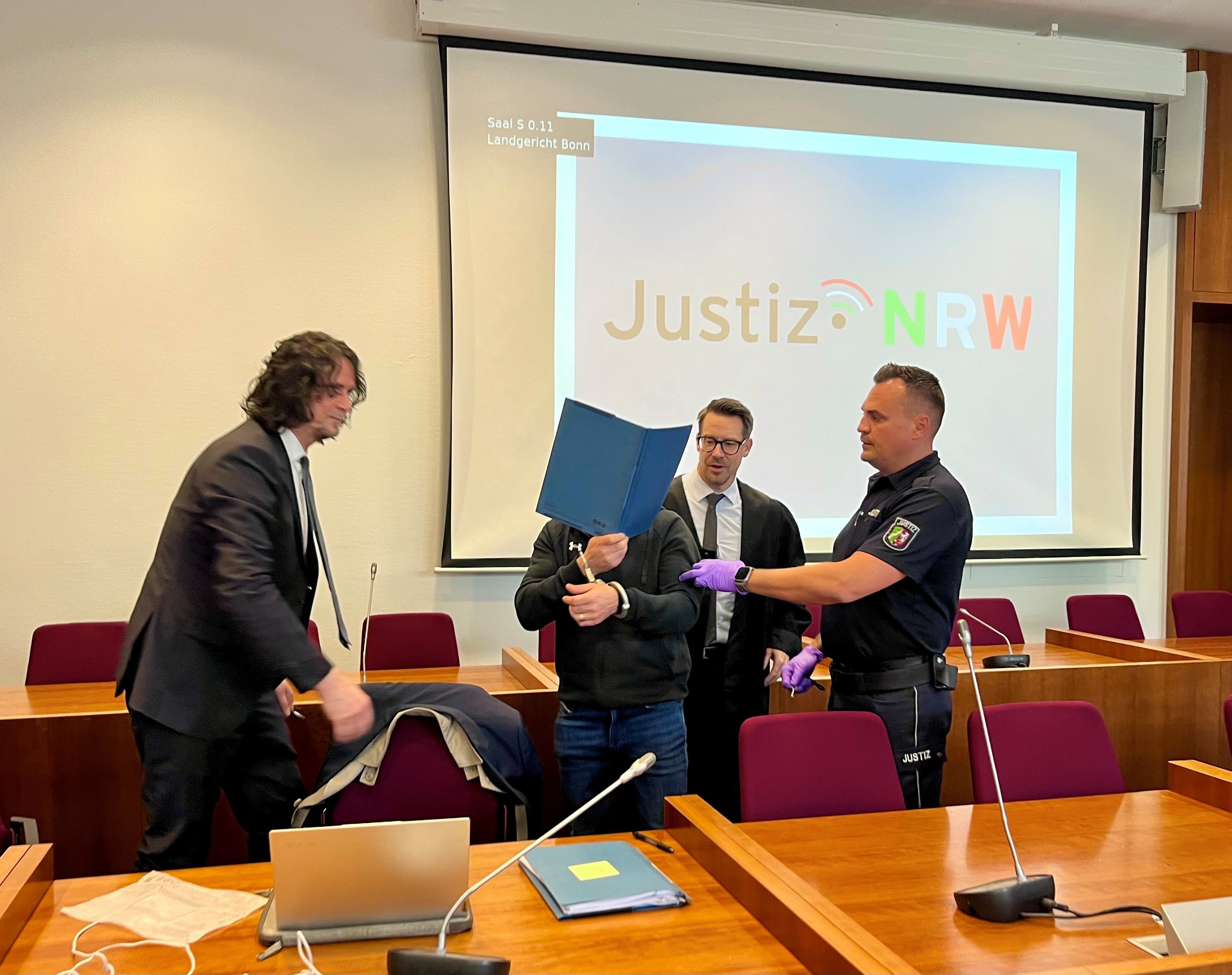 Ein Justizmitarbeiter führt den Angeklagten, der sich eine blaue Mappe vors Gesicht hält. Rechts und links von ihm steht jeweils ein Verteidiger im dunklen Anzug.
