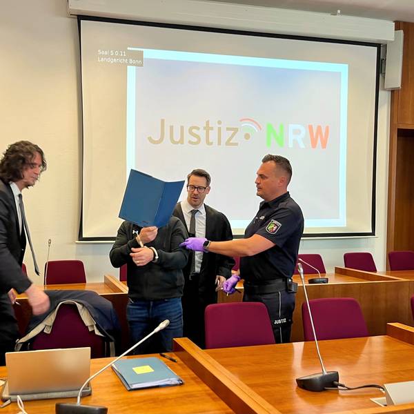 Ein Justizmitarbeiter führt den Angeklagten, der sich eine blaue Mappe vors Gesicht hält. Rechts und links von ihm steht jeweils ein Verteidiger im dunklen Anzug.