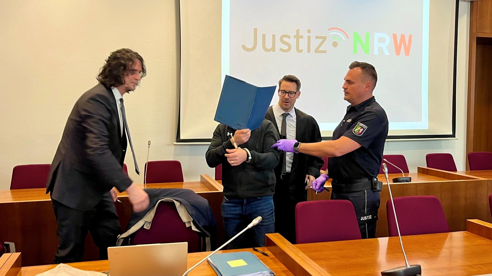 Ein Justizmitarbeiter führt den Angeklagten, der sich eine blaue Mappe vors Gesicht hält. Rechts und links von ihm steht jeweils ein Verteidiger im dunklen Anzug.