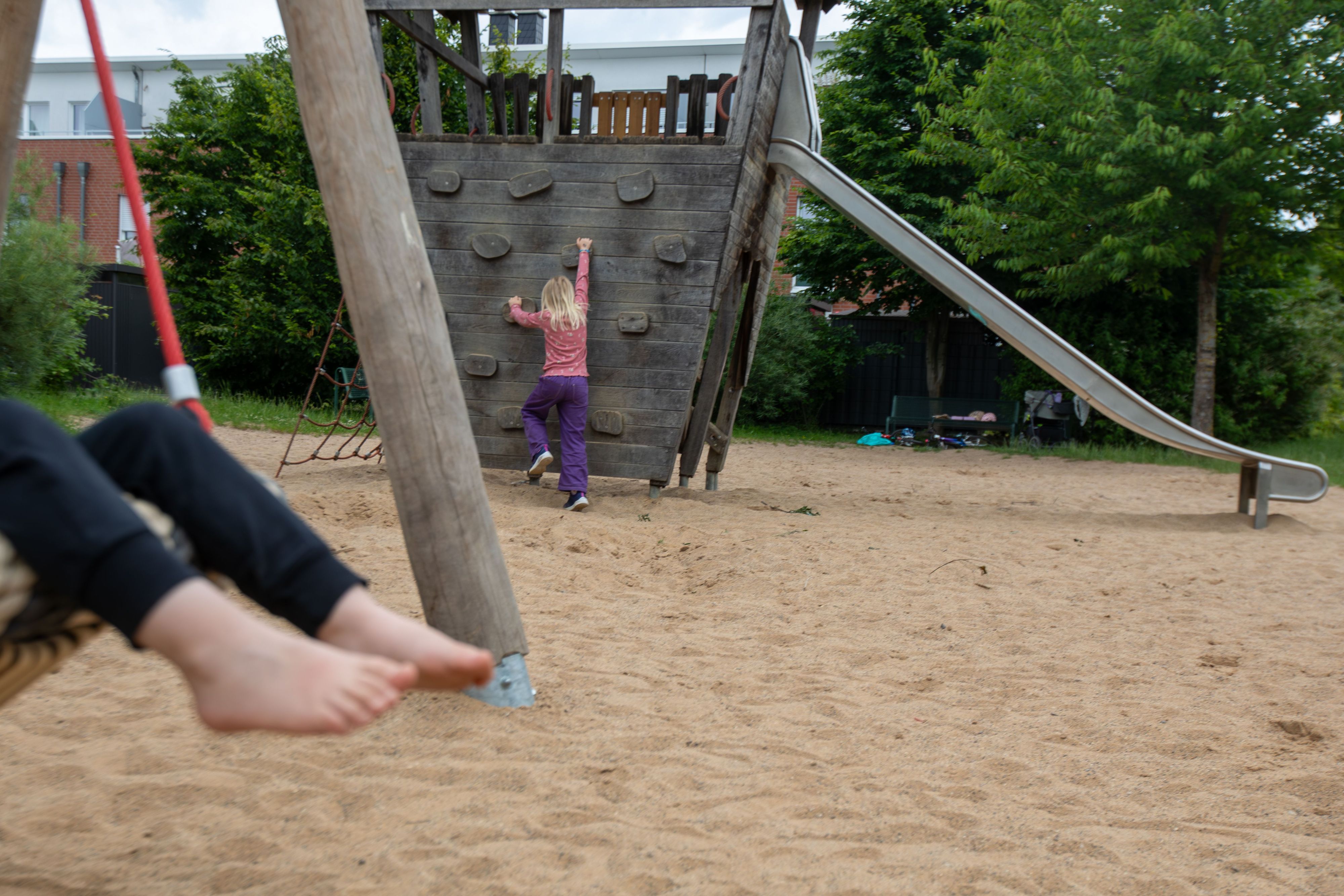 Auf einem Spielplatz ist ein schaukelndes Kind zu sehen. Im Hintergrund klettert ein Mädchen.