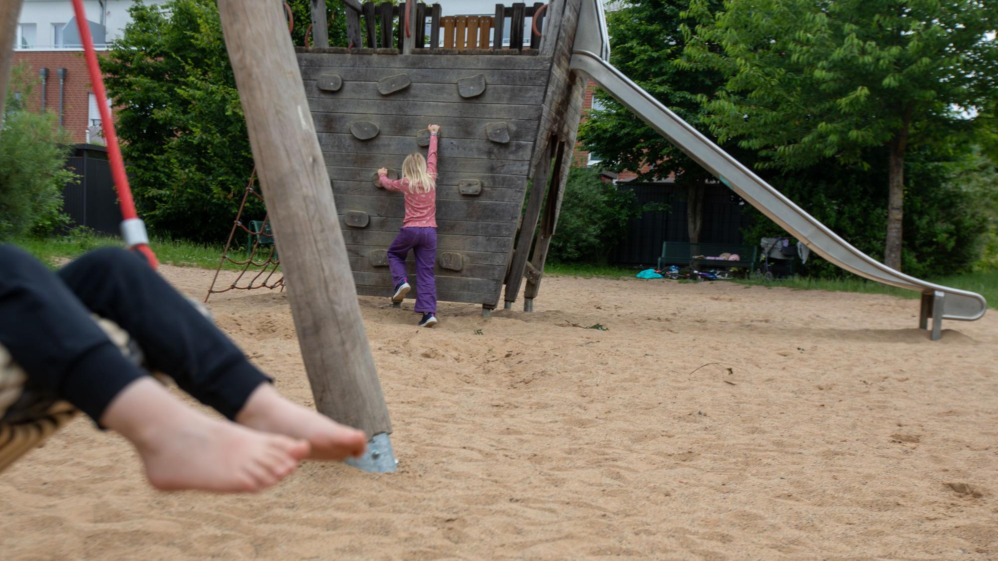 Auf einem Spielplatz ist ein schaukelndes Kind zu sehen. Im Hintergrund klettert ein Mädchen.