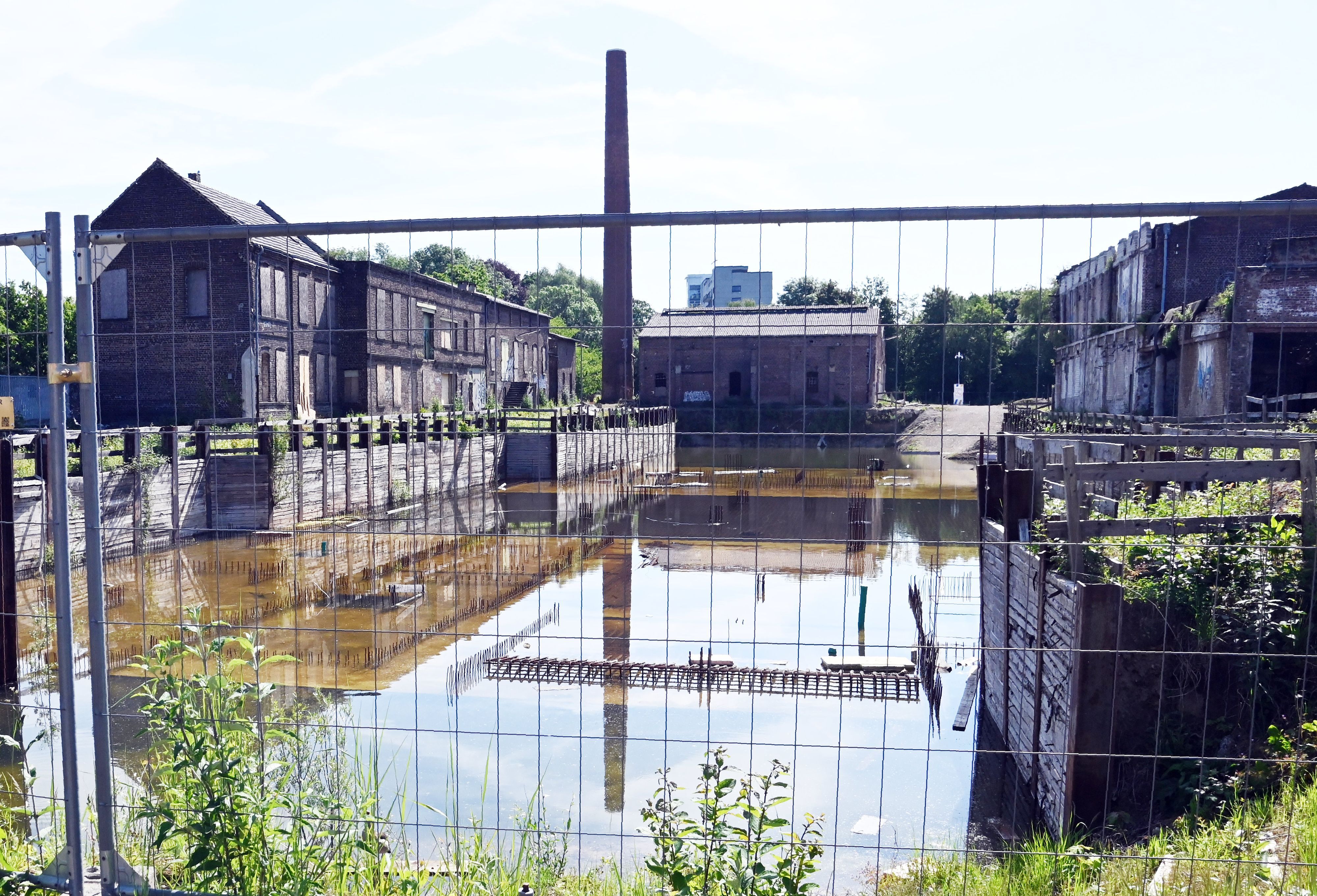 Ein altes Industriegelände mit Backstein-Fabrikgebäuden und Schornstein liegt hinter einem Bauzaun.