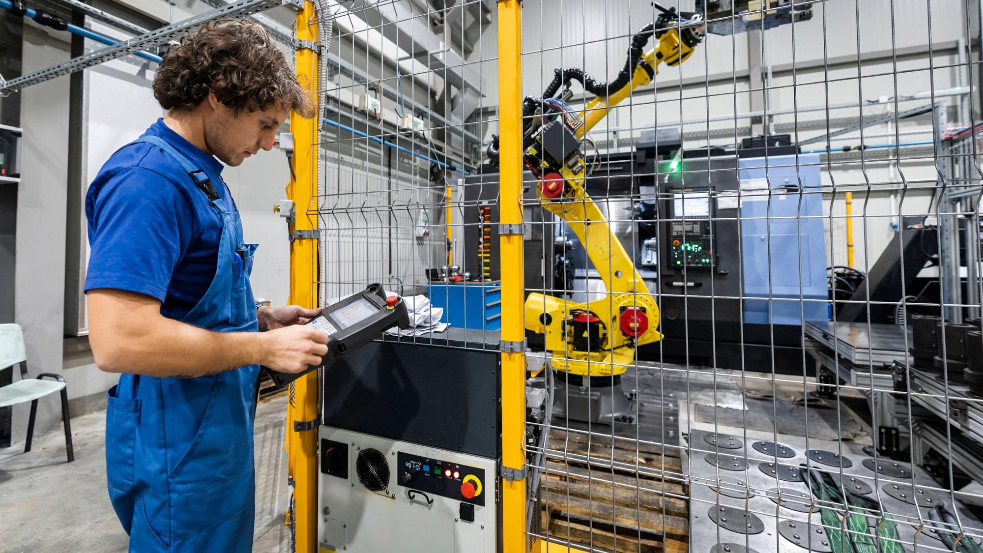 Ein Mann im Blaumann steht in einer Produktionshalle an einer Maschine mit Roboterarm.
