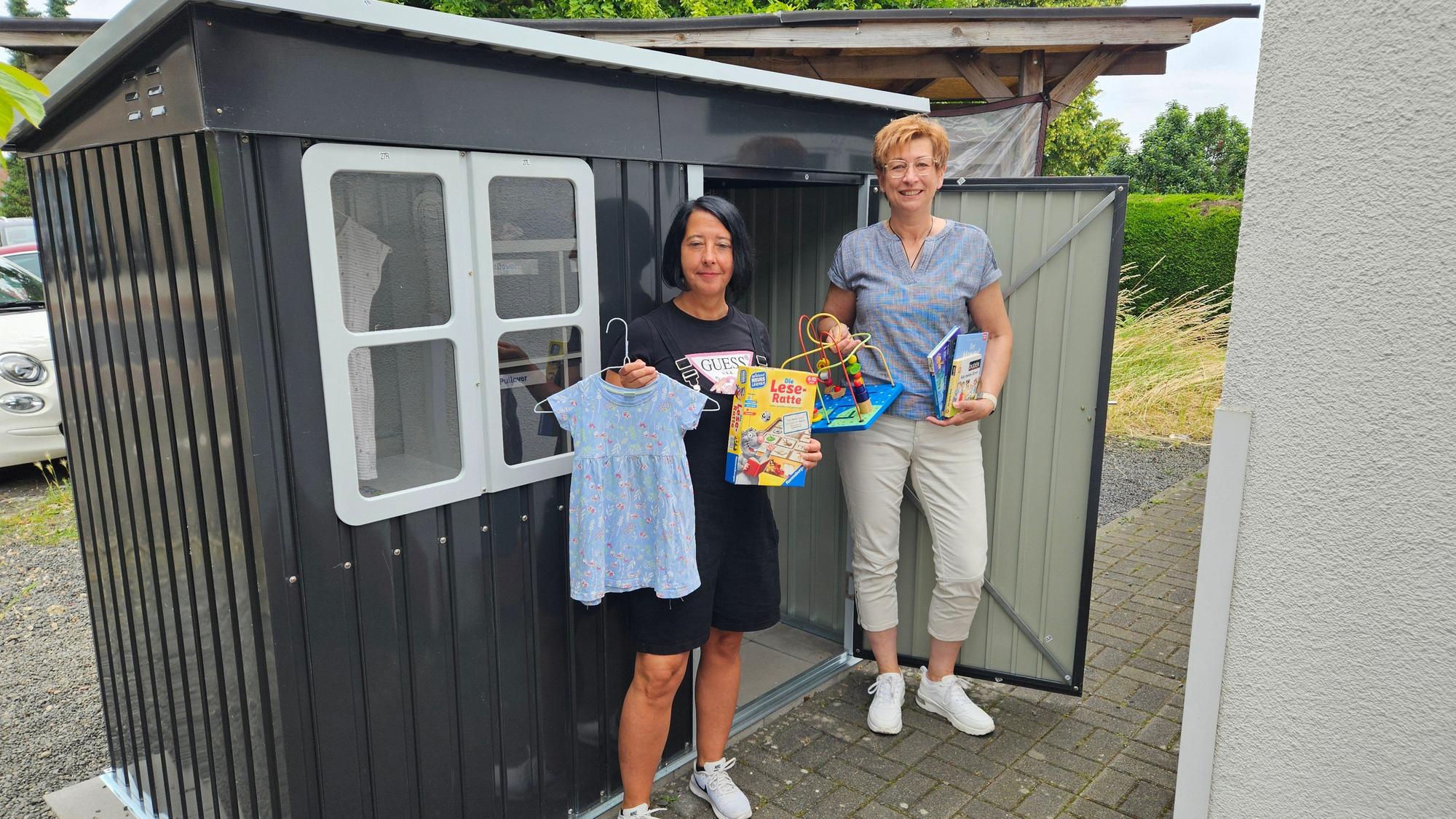 Zu sehen sind zwei Frauen mit Spielzeug und Kleidung für Kinder vor einem Gartenhäuschen.