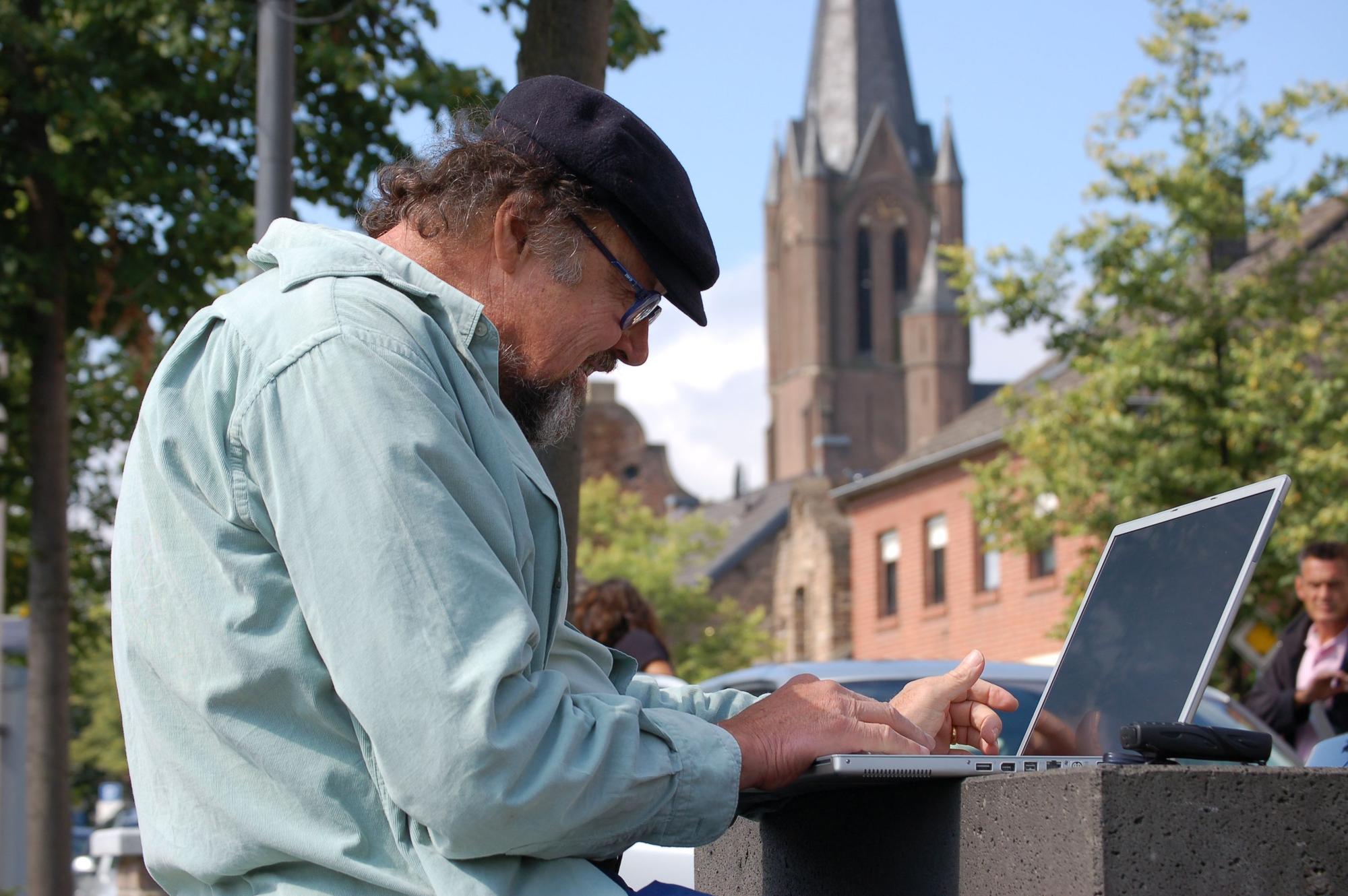 Ein Mann schaut auf ein vor ihm stehendes Laptop, im Hintergrund eine Kirche.