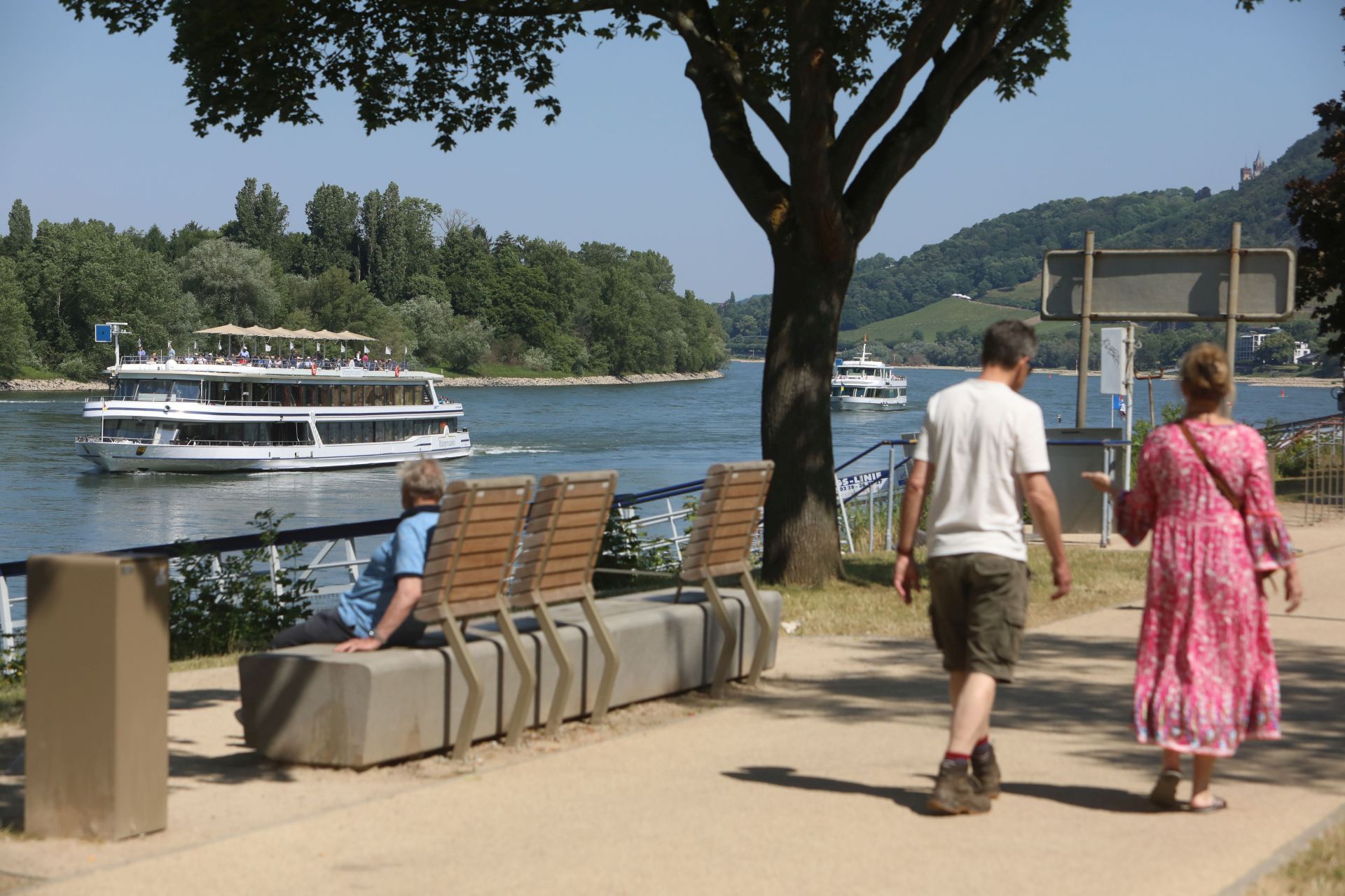 Ein Paar geht über die Insel, links sitzt eine Person auf einer Bank und blickt auf ein vorbeifahrendes Ausflugsschiff.