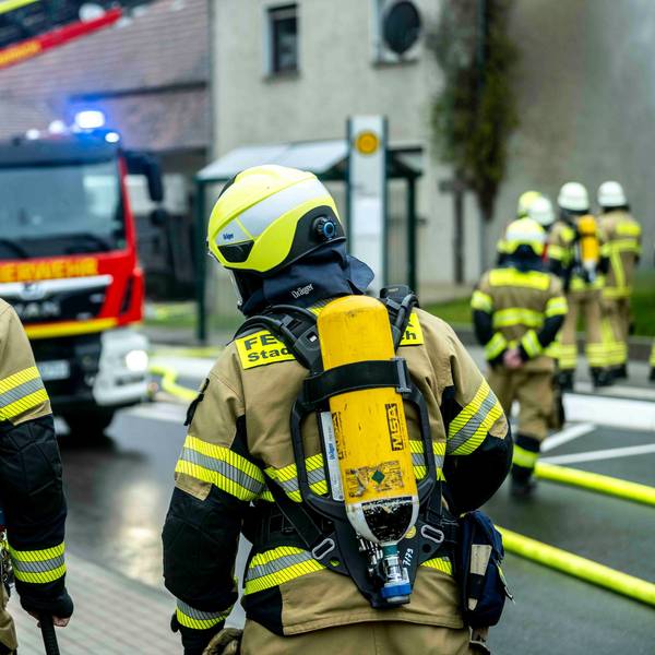 Zwei Feuerleute mit Pressluftatmern auf dem Rücken schauen auf eine Einsatzstelle, an der auch eine Drehleiter steht.
