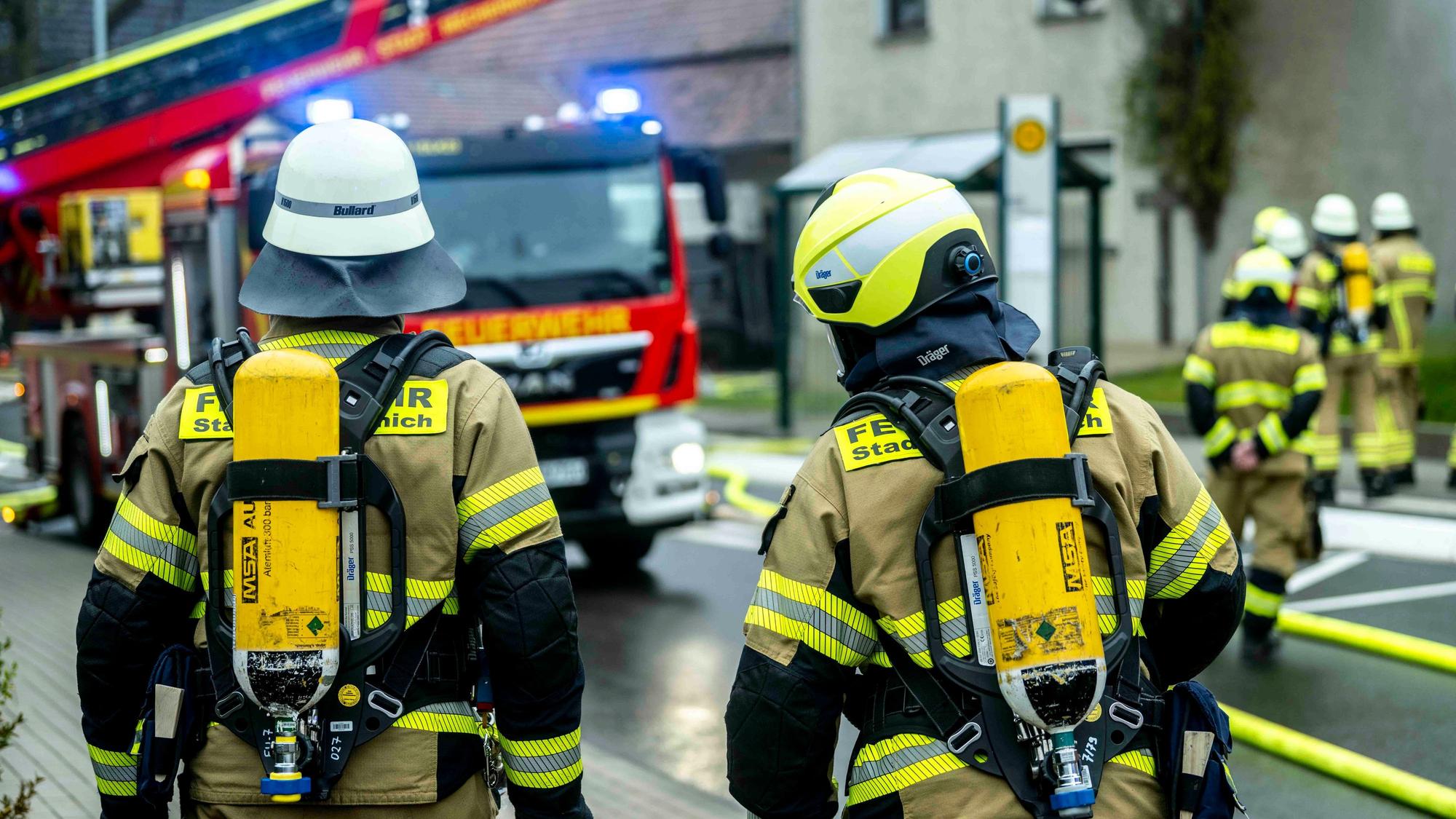 Zwei Feuerleute mit Pressluftatmern auf dem Rücken schauen auf eine Einsatzstelle, an der auch eine Drehleiter steht.