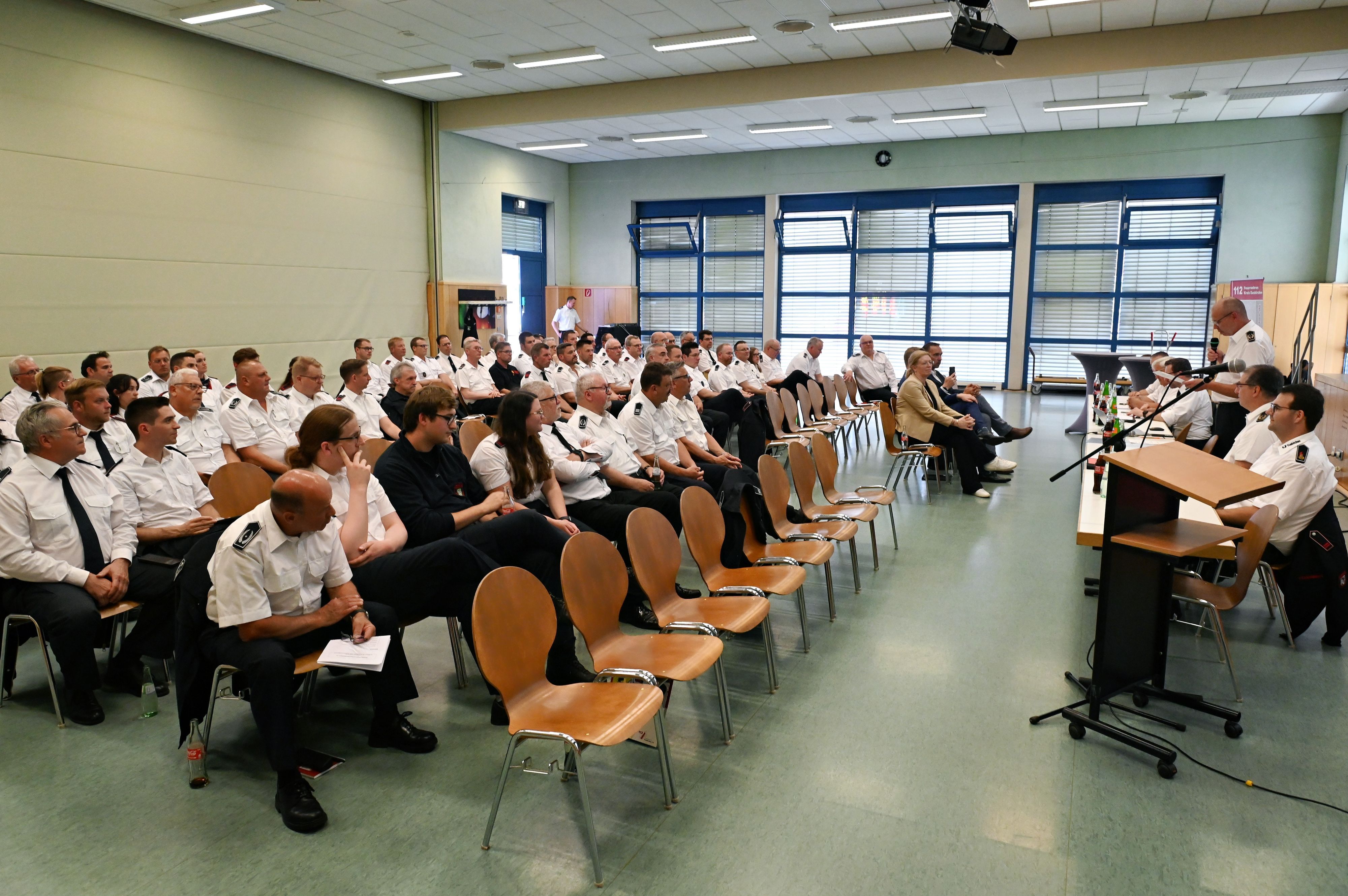 Zahlreiche Feuerwehrleute in weißen Uniform-Hemden sitzen in einem Saal.