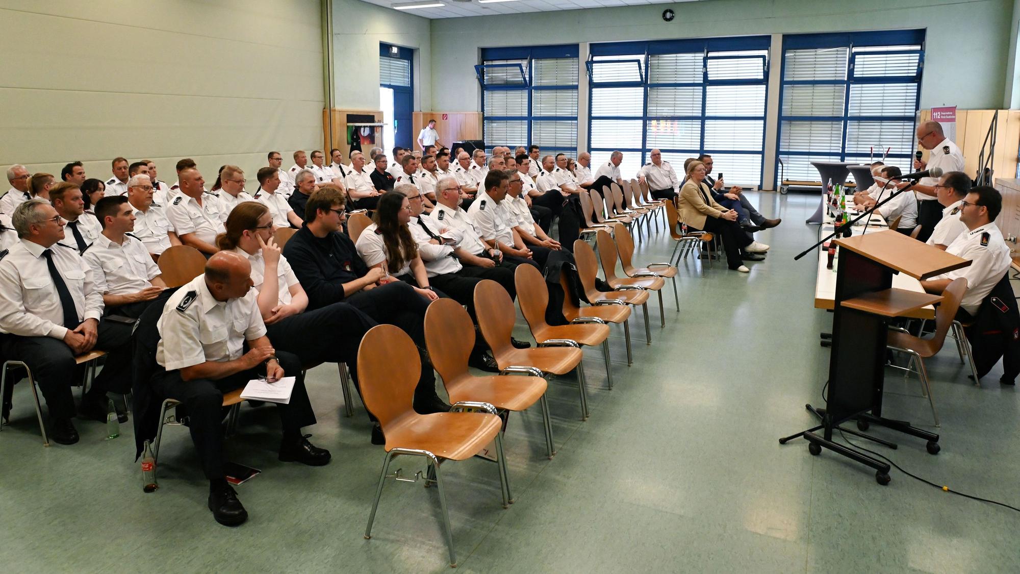 Zahlreiche Feuerwehrleute in weißen Uniform-Hemden sitzen in einem Saal.