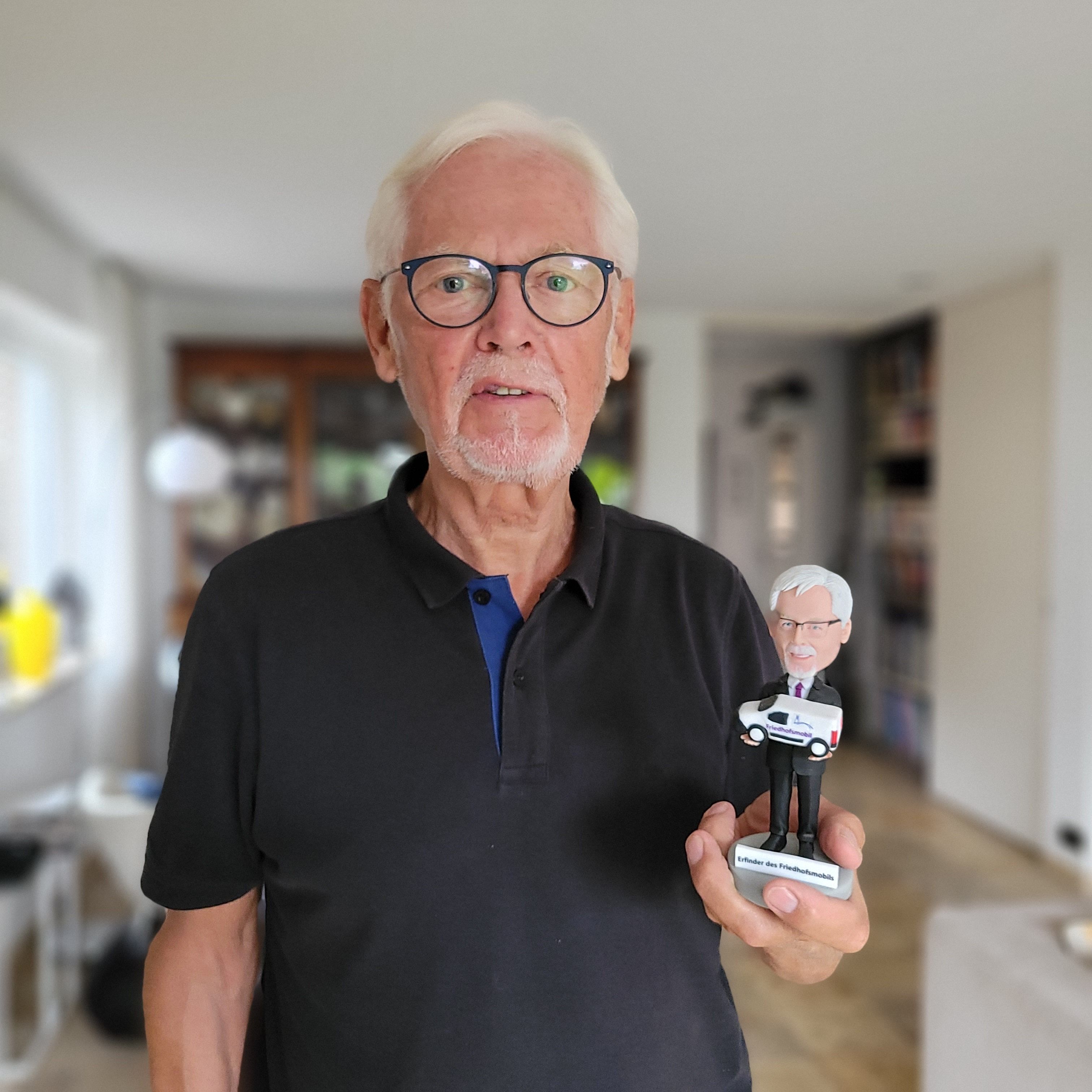 Ein Mann mit weißem Bart und weißen Haaren hält eine Figur in der Hand. Sie stellt ihn selbst dar.