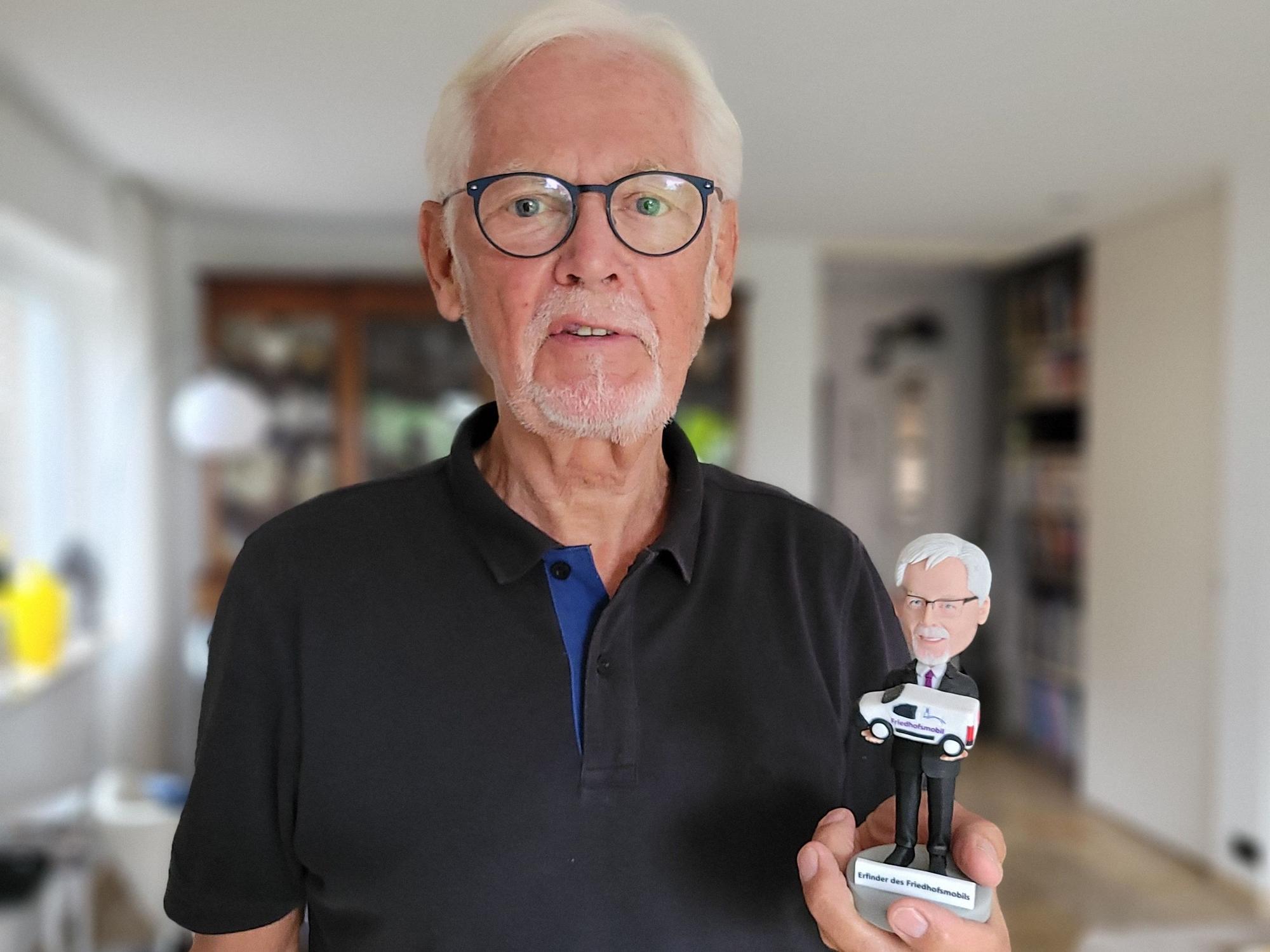 Ein Mann mit weißem Bart und weißen Haaren hält eine Figur in der Hand. Sie stellt ihn selbst dar.