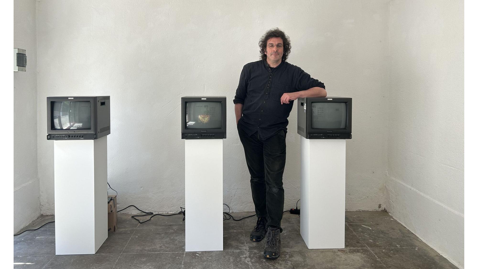 Tobias Schmücking vor seiner Videoarbeit mit drei japanischen Winkekatzen, die fragmentarisch auf alten Studiomonitoren zu sehen sind.