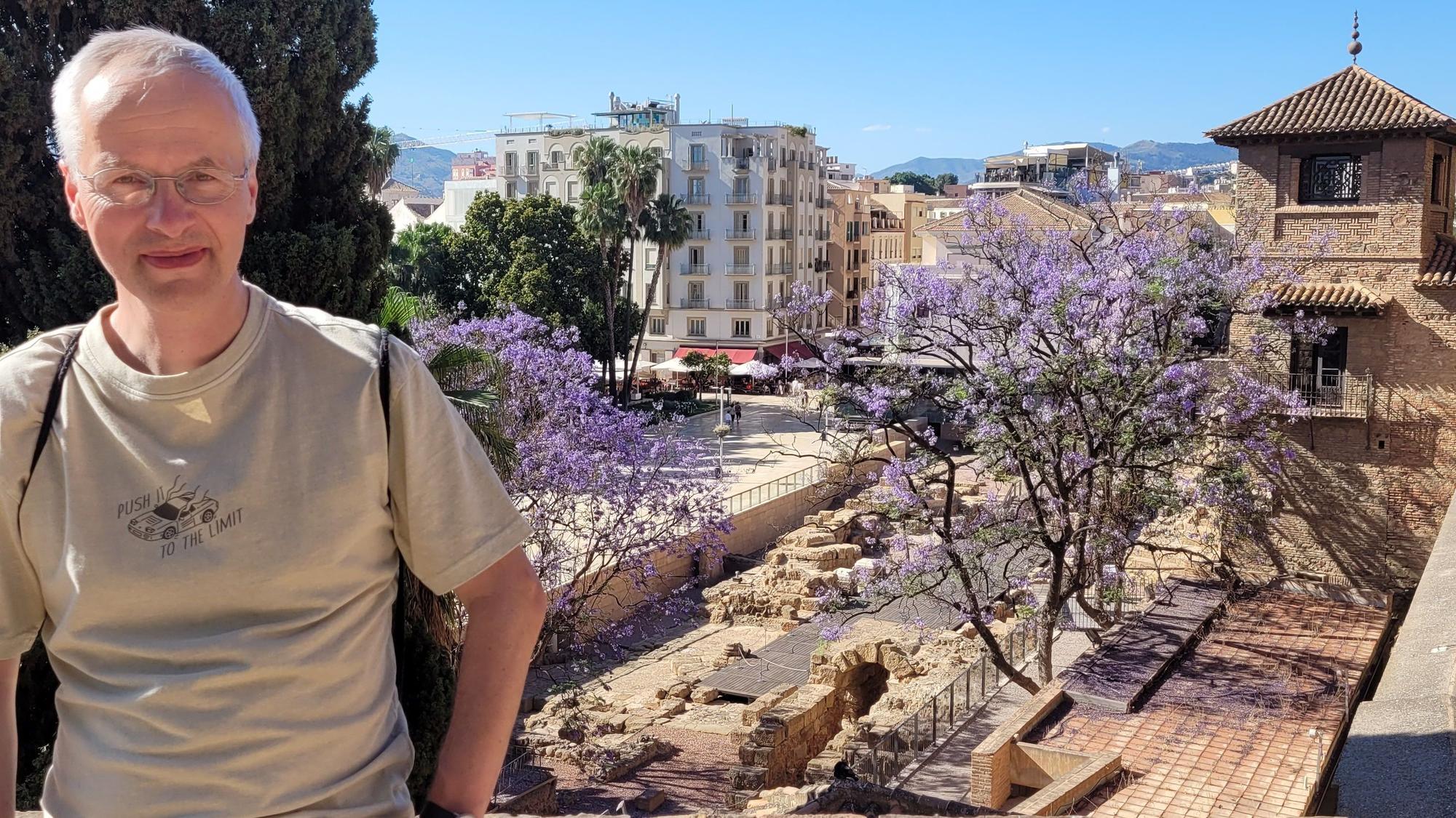 Zu sehen ist ein Mann in einer mediterranen Stadt, der in die Kamera lächelt.