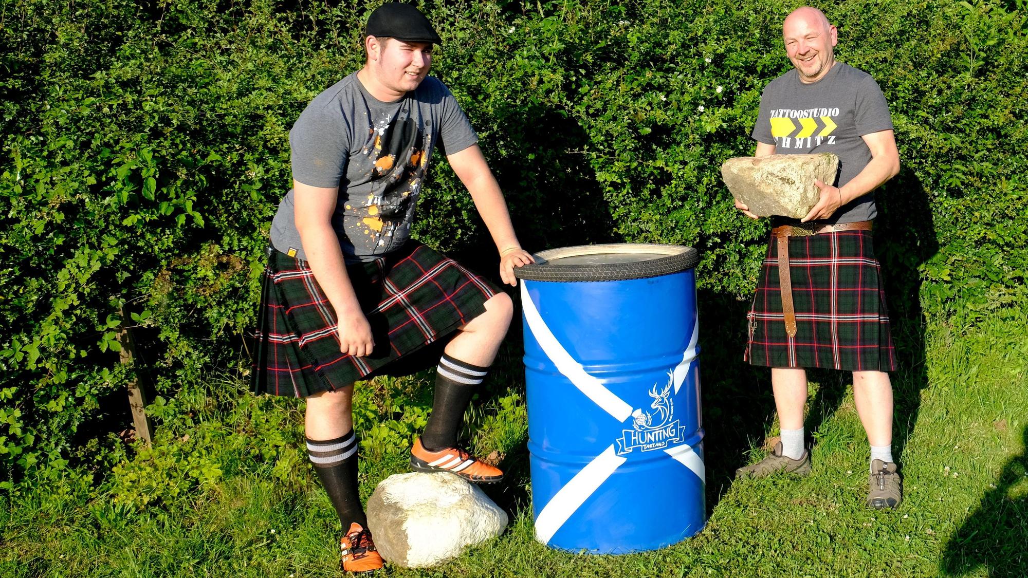 Der Redakteur im Schottenrock trägt einen Stein zu einem blauen Metallfass, daneben steht Louis Hochgürtel, ebenfalls im Kilt, der einen Fuß auf einen großen Stein gestellt hat.