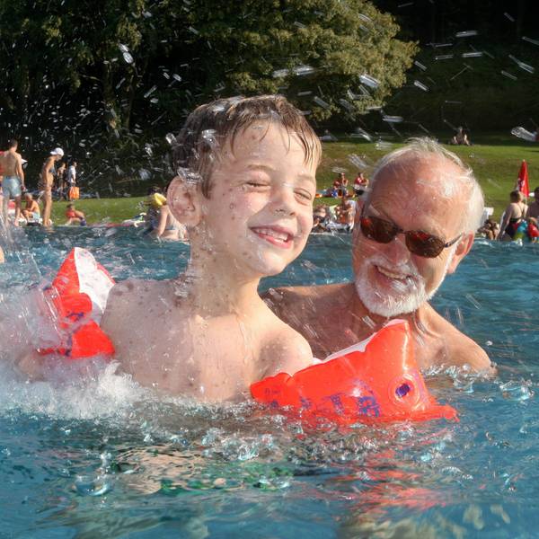 Ein Mann kühlt sich mit einem Kind mit Schwimmflügeln unter einem Wasserschwall im Nichtschwimmerbecken des Freibads Hoffnungsthal ab.