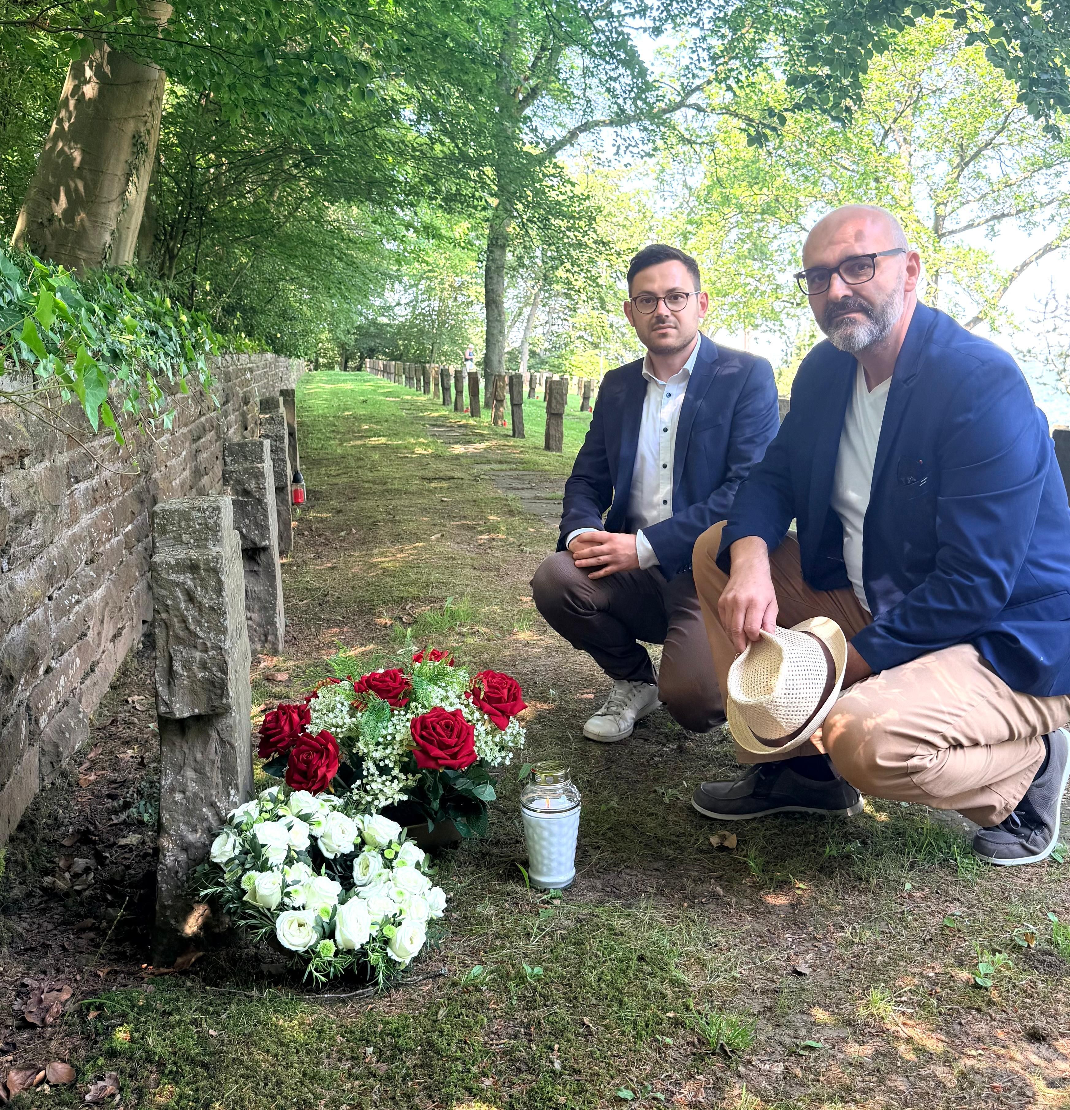 Zwei Männer knien vor einem Grab, auf dem Blumen stehen.