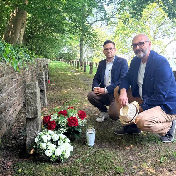 Zwei Männer knien vor einem Grab, auf dem Blumen stehen.
