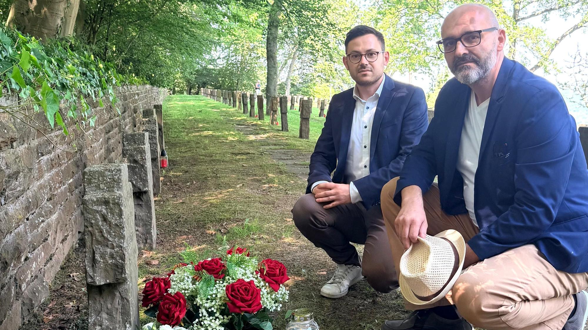 Zwei Männer knien vor einem Grab, auf dem Blumen stehen.