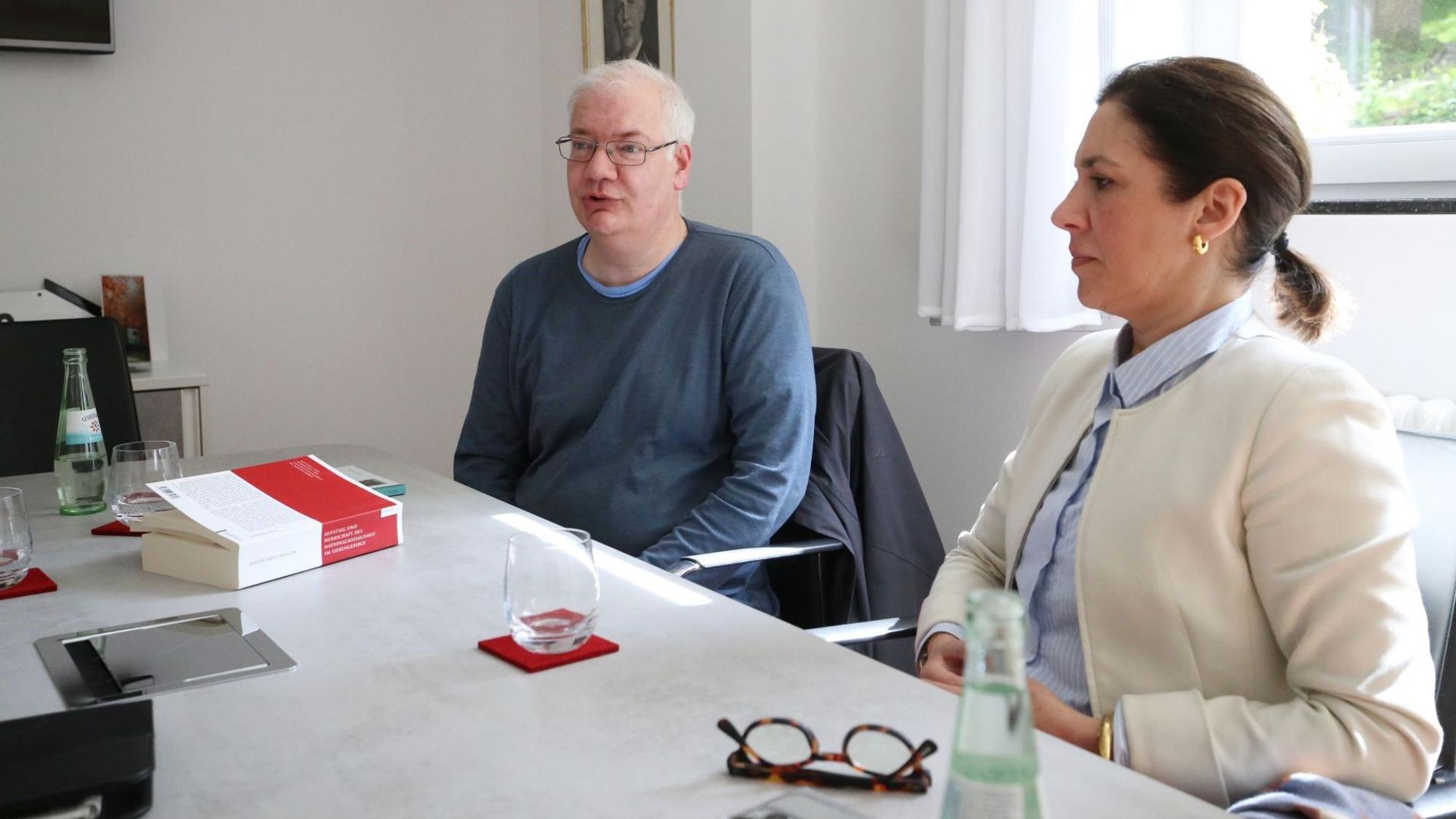 Die Obengenannten sitzen an einem Tisch und schauen in unterschiedliche Richtungen auf etwas außerhalb des Bildausschnitts.