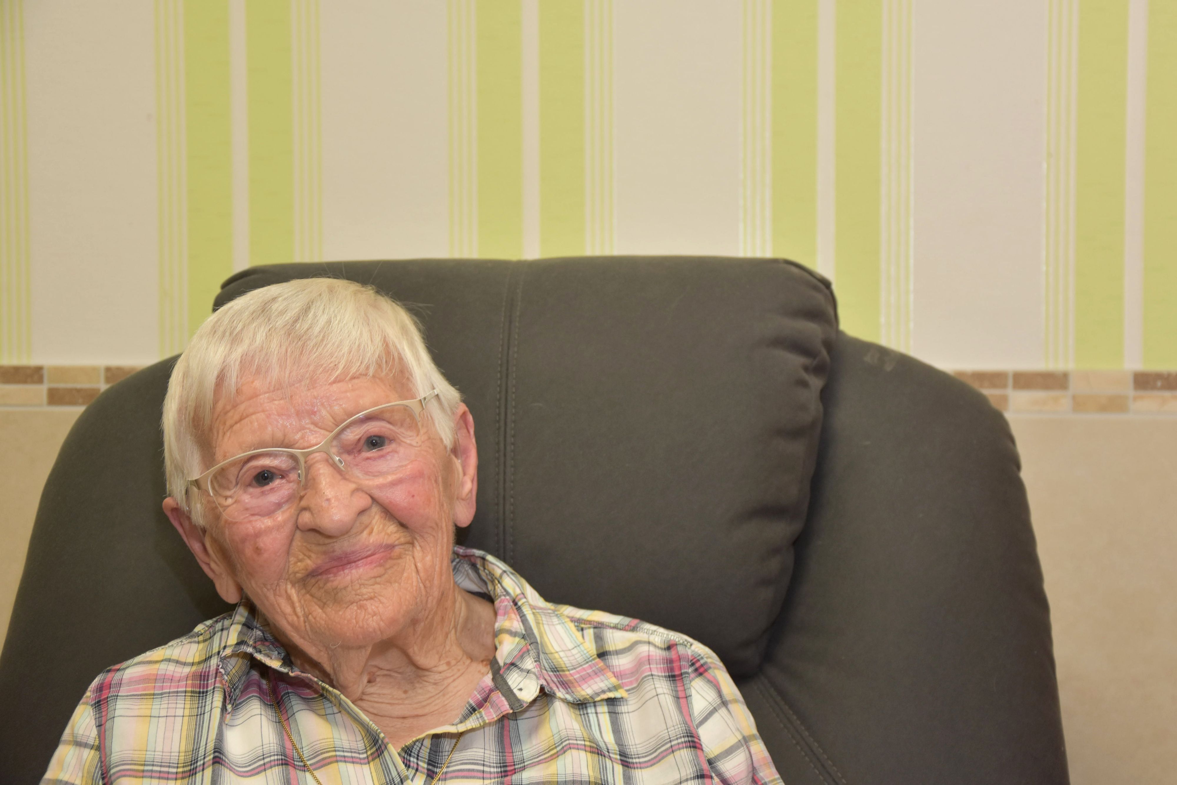 Das Bild zeigt die Seniorin auf einem Sofa. Sie trägt ein kariertes Hemd.