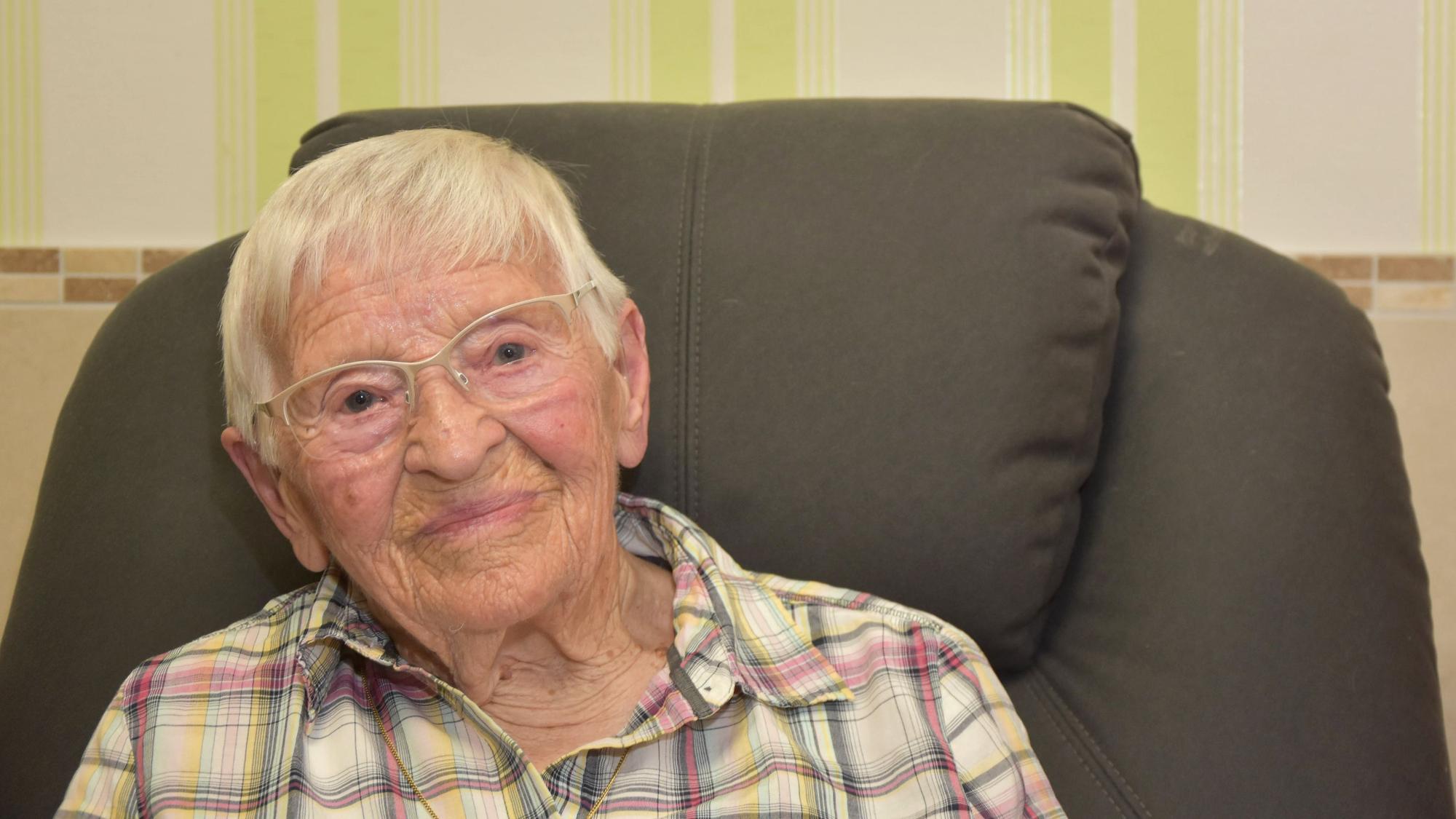 Das Bild zeigt die Seniorin auf einem Sofa. Sie trägt ein kariertes Hemd.