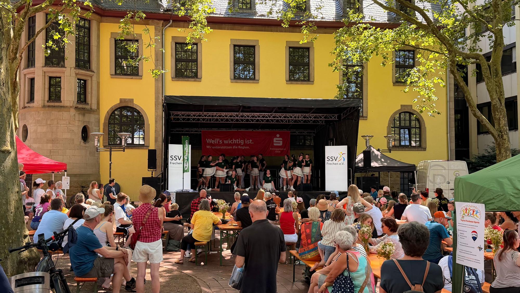 Das Foto zeigt Gäste eines Sportfests, im Hintergrund findet eine Bühnenaufführung statt.