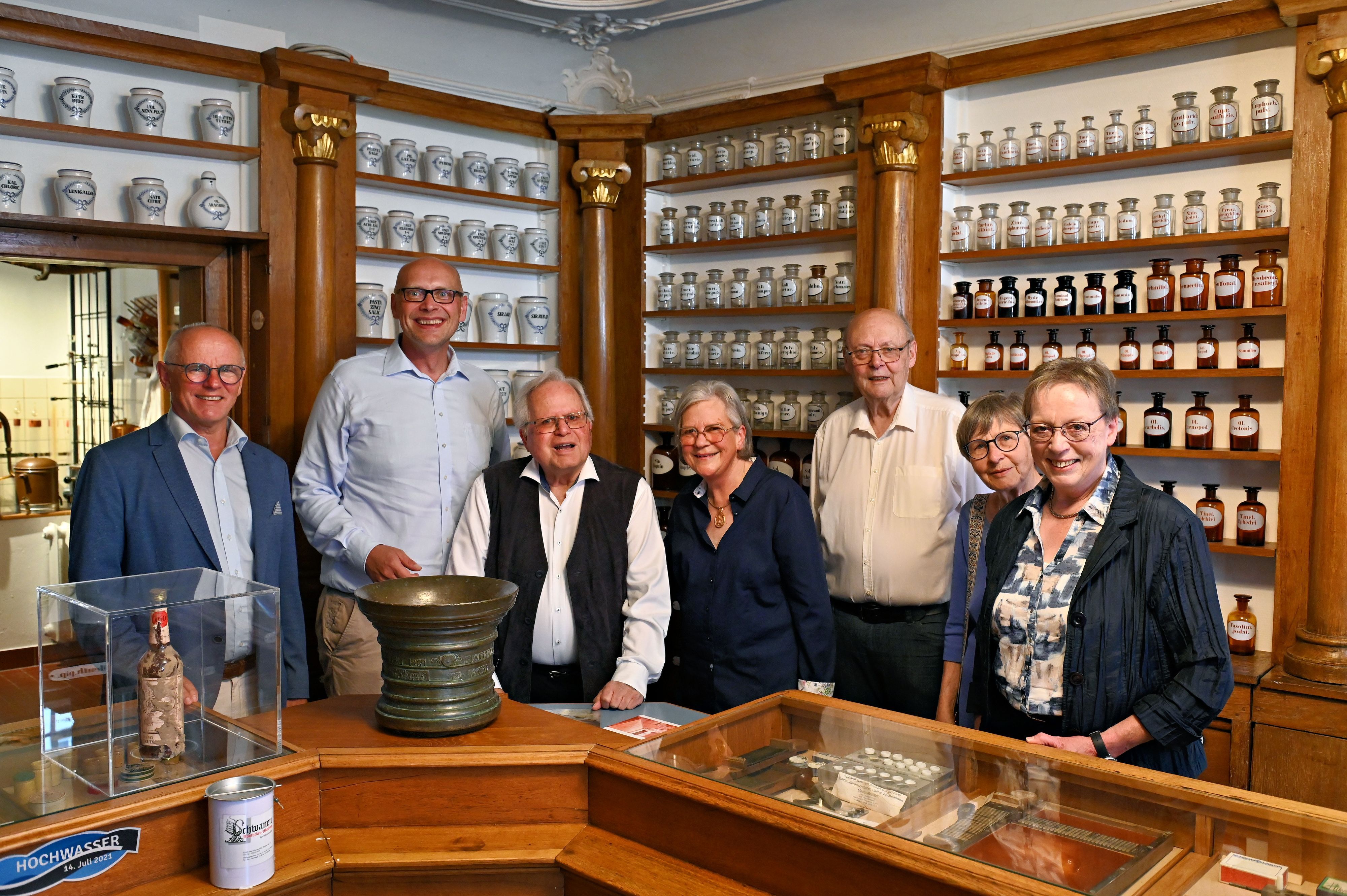 Die genannten Personen stehen im historischen Apothekengebäude vor Regalen mit zahlreichen Flaschen und anderen Behältnissen.