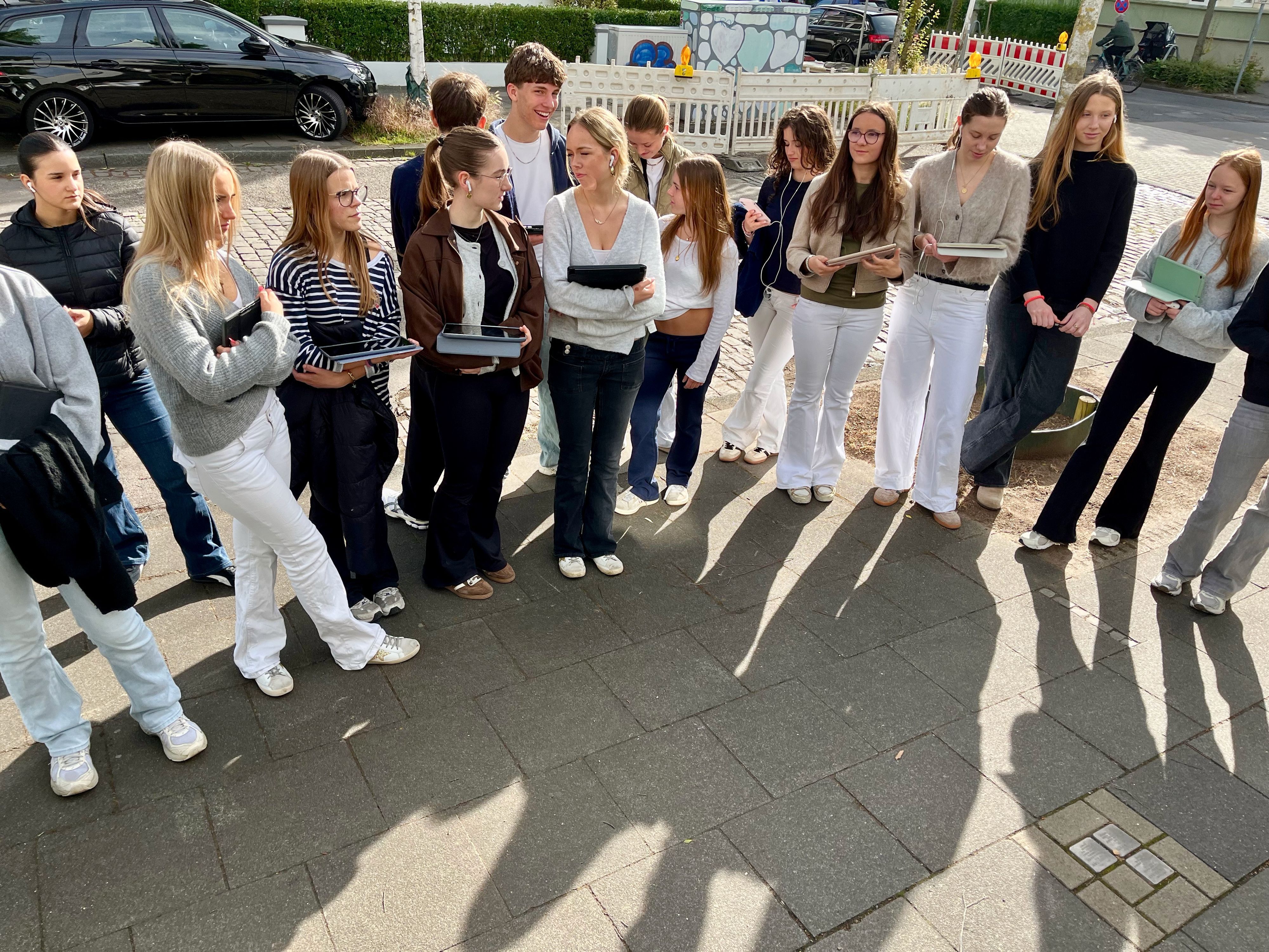 Schülerinnen und Schüler stehen nebeneinander vor drei Stolpersteinen auf einem Bürgersteig an einer Straßenkreuzung.