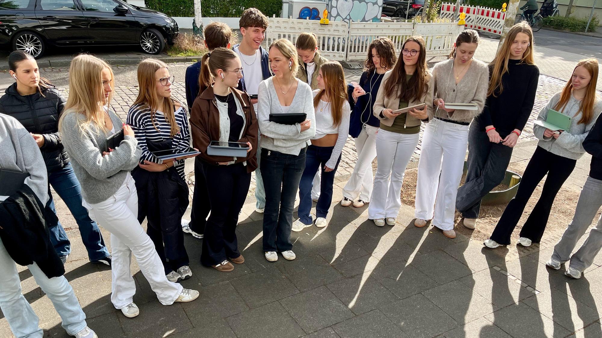 Schülerinnen und Schüler stehen nebeneinander vor drei Stolpersteinen auf einem Bürgersteig an einer Straßenkreuzung.