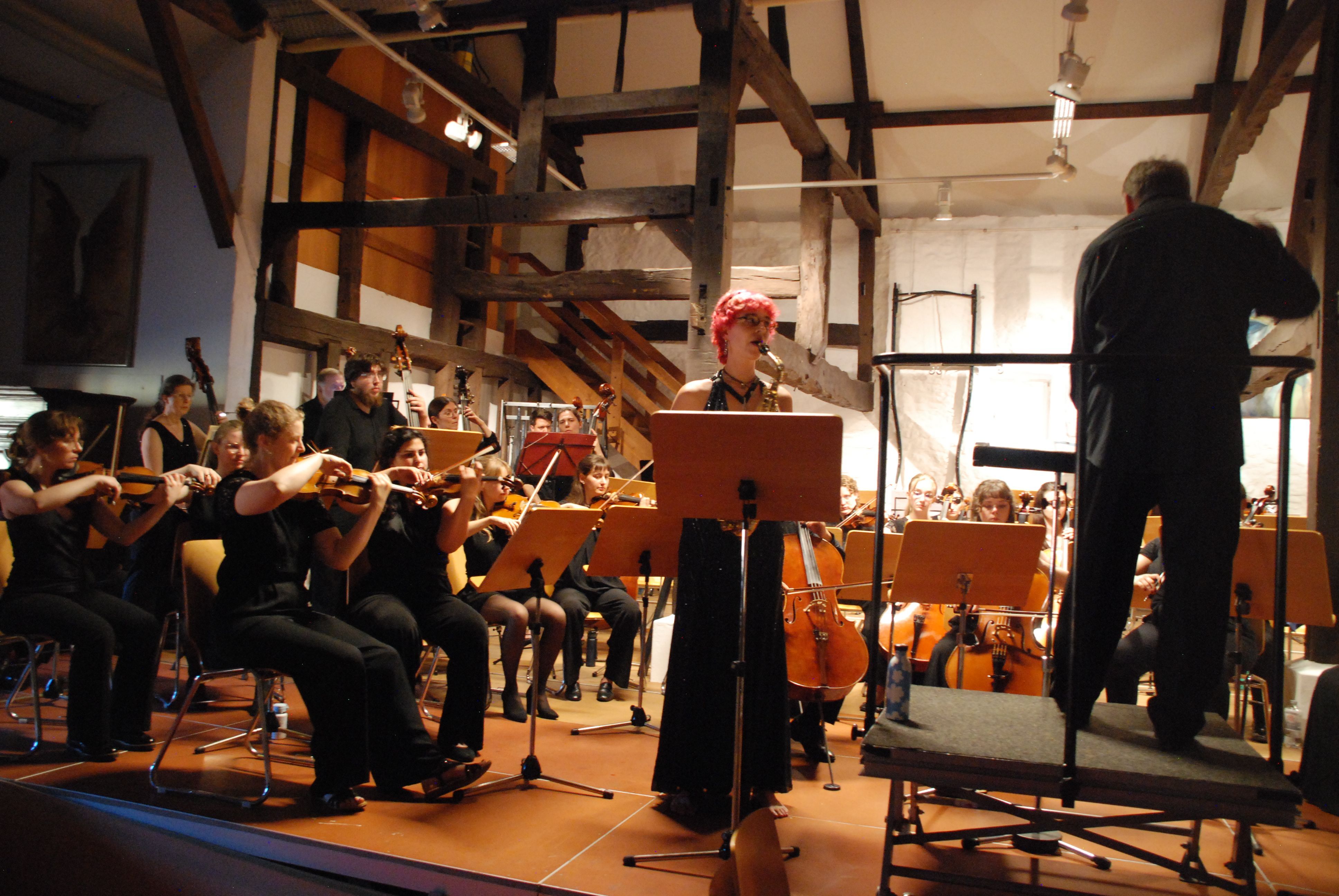 Lilian Eicks steht hinter einem Notenpult und spielt Saxofon. Links von ihr befindet sich das Orchester, rechts der Dirigent,.